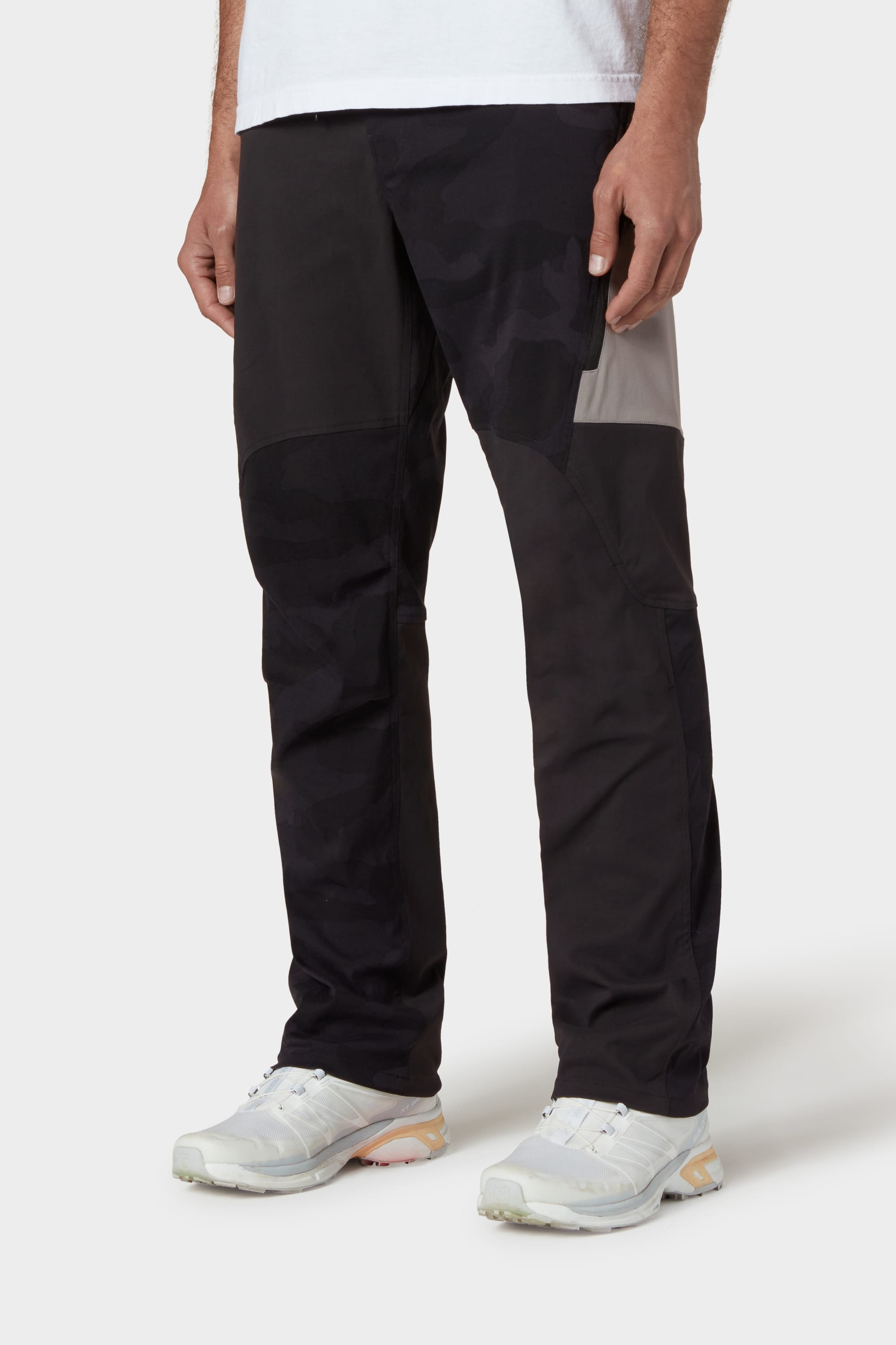 A person wearing the 686 Mens Anything Cargo Pant - Relaxed Fit in black and gray patchwork, paired with a white shirt and white sneakers, stands against a plain light background.
