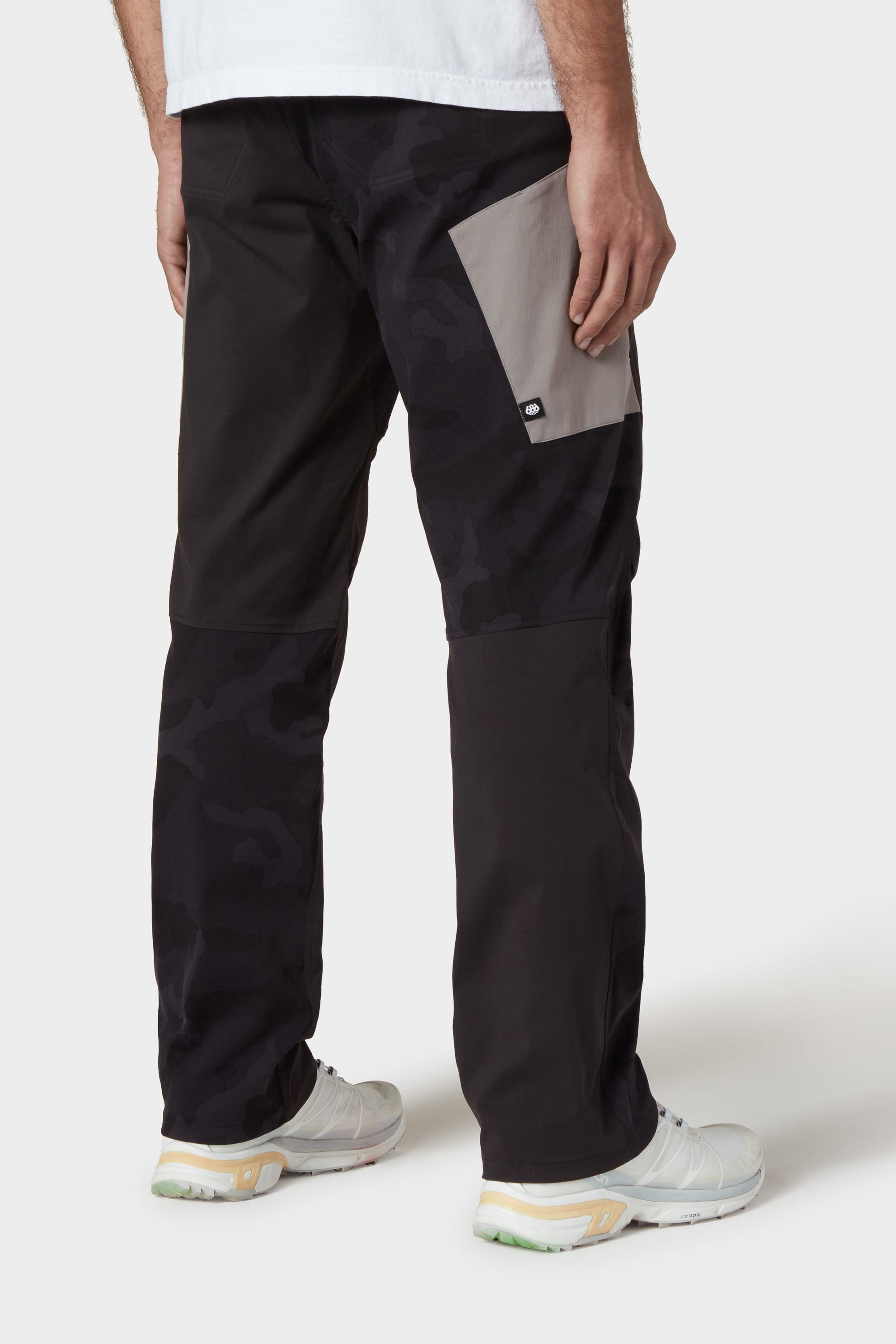 A person in 686 Mens Anything Cargo Pant - Relaxed Fit by 686, featuring black and dark camo four-way stretch, a light gray back pocket, styled with white sneakers and a white shirt, stands against a plain white background.