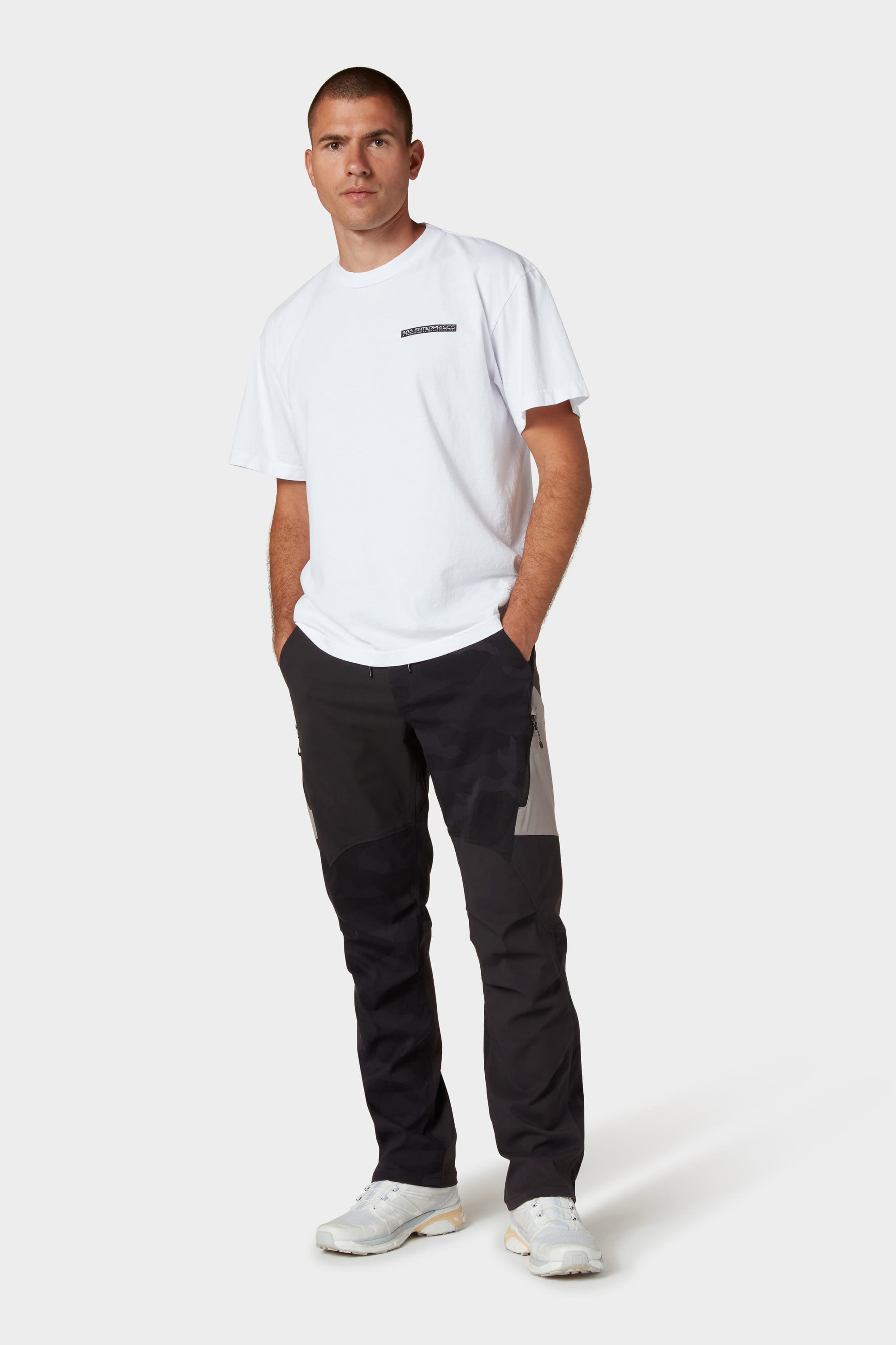 A person stands against a plain background wearing a white T-shirt, 686 Mens Anything Cargo Pant - Relaxed Fit in black and gray by 686, and white sneakers, with hands in pockets and a neutral expression.