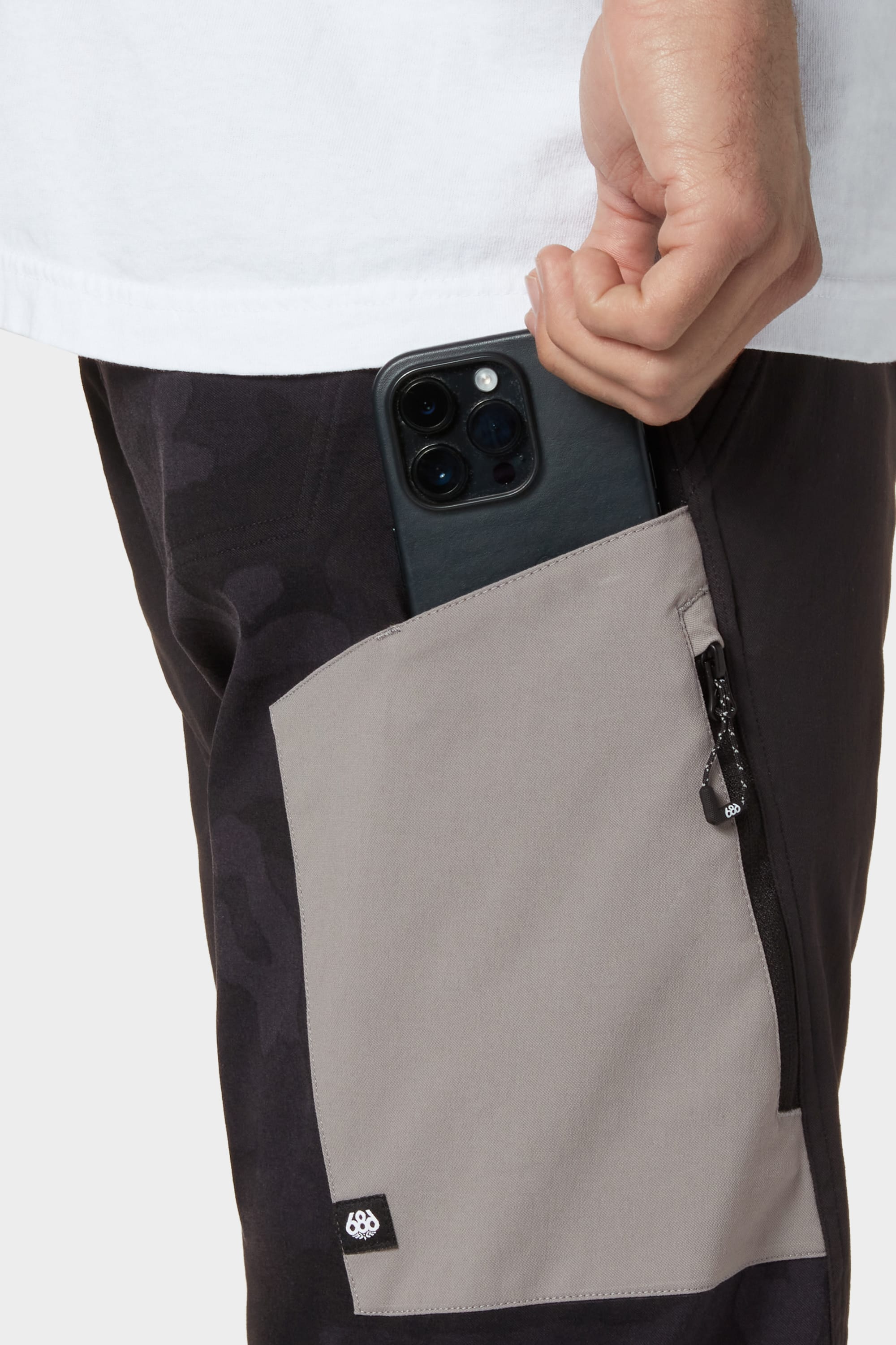 A person is sliding a black smartphone into the zippered side pocket of 686 Mens Anything Cargo Pant - Relaxed Fit by 686, in two-tone black and gray with breathable, 4-way stretch fabric, while wearing a white shirt.