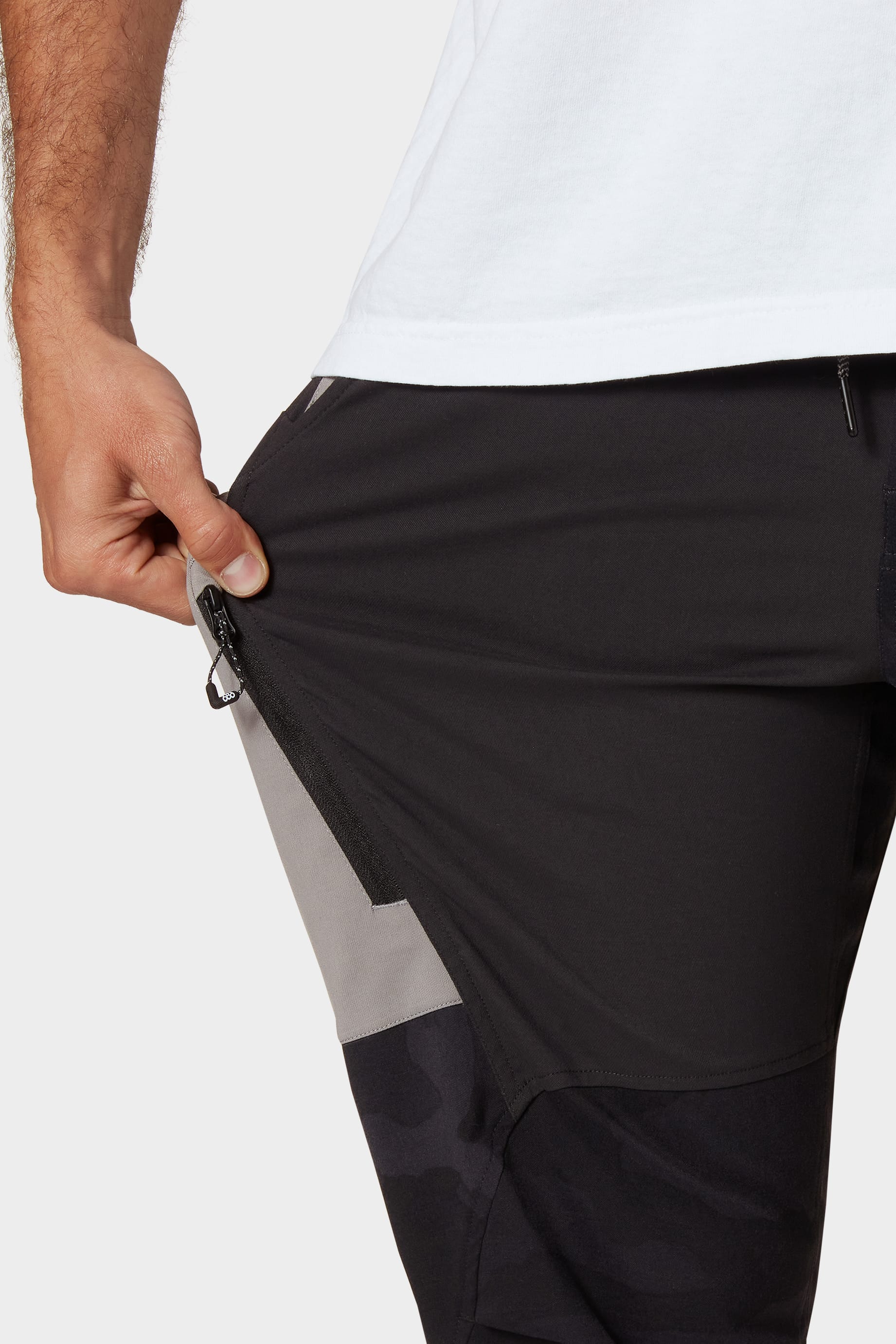 A person wearing 686 Mens Anything Cargo Pant - Relaxed Fit in black and gray stretches the 4-way stretch fabric near the hip, highlighting its flexibility. Only their torso, arm, hand, and a white shirt are visible. Brand: 686.