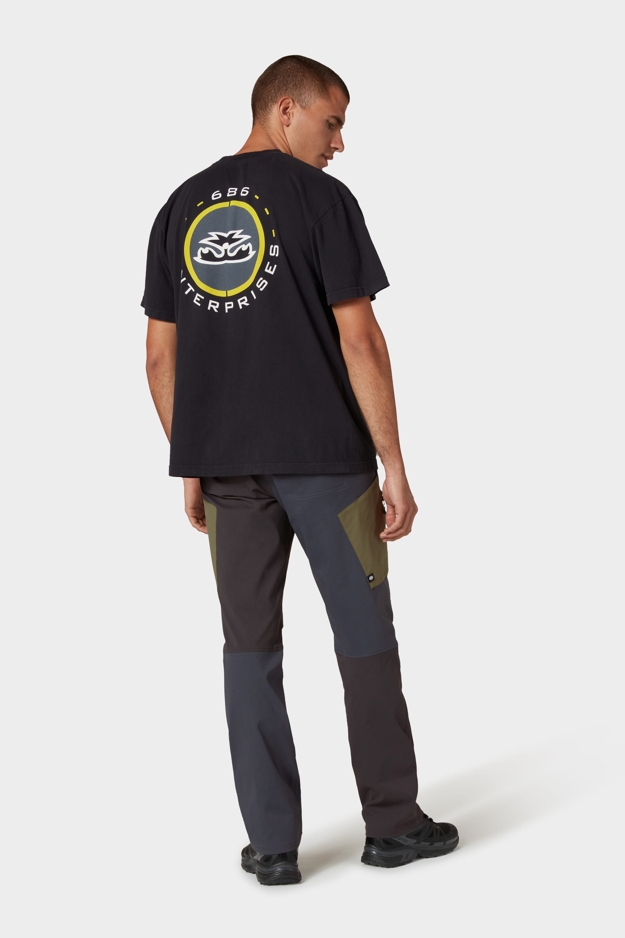 A person stands facing slightly away, wearing a black T-shirt with a circular graphic and text on the back, 686 Mens Anything Cargo Pant - Relaxed Fit by 686 in dark and olive green, and black shoes against a plain light background.