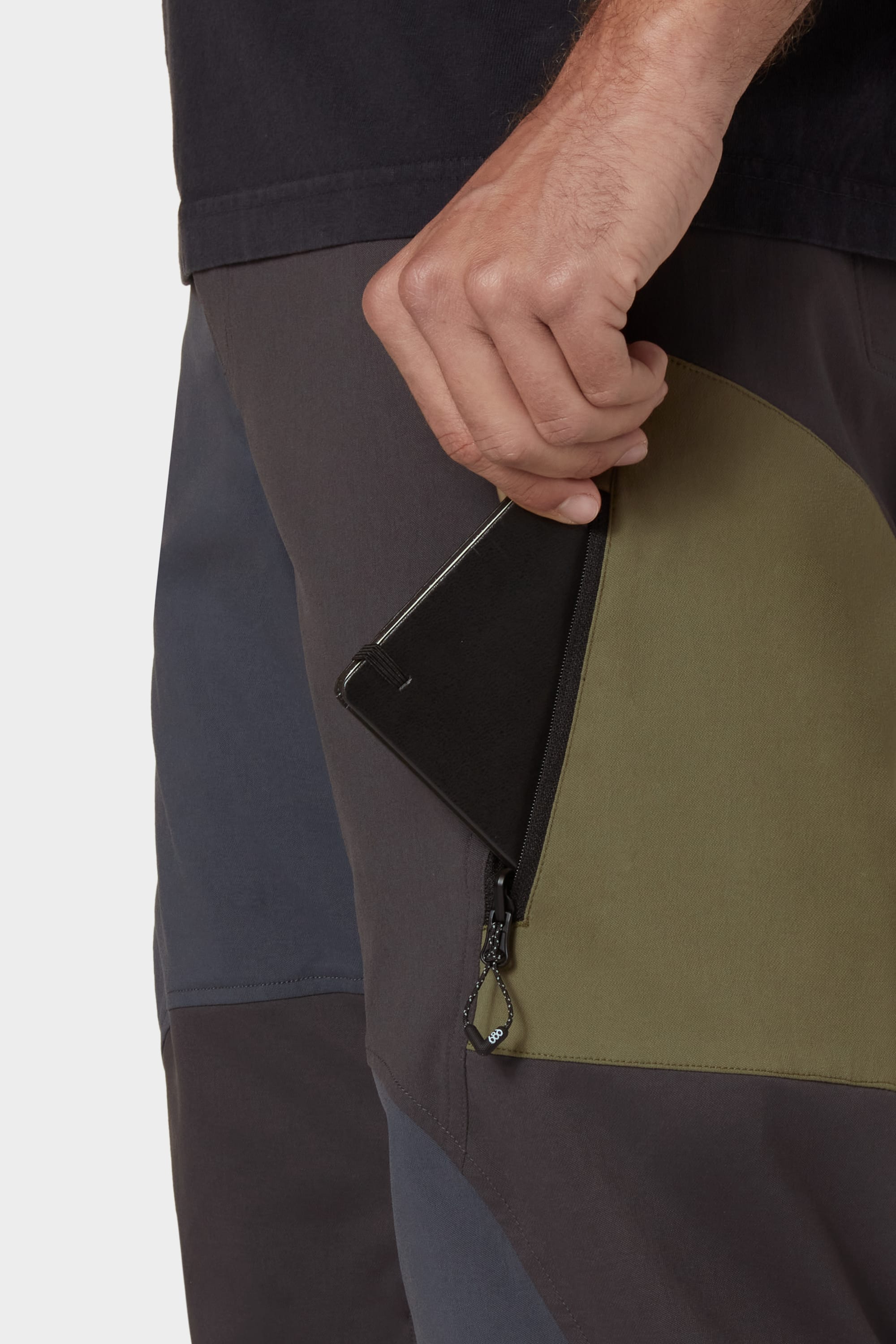 A person slides a black smartphone into the zippered side pocket of the 686 Mens Anything Cargo Pant - Relaxed Fit by 686, featuring olive green and dark gray breathable panels.