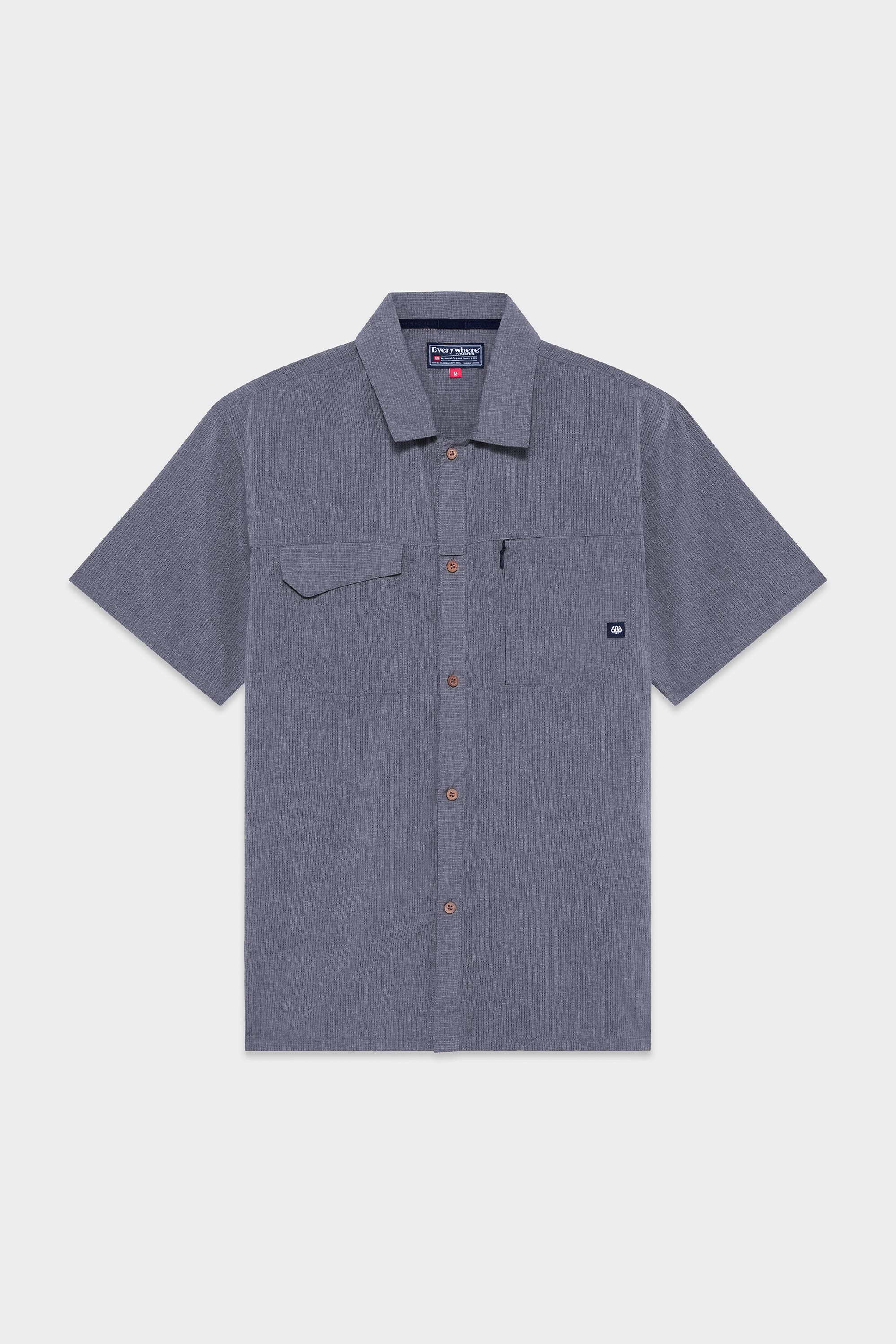 The 686 Mens Seeker Perforated Button Up features short sleeves, a collar, brown buttons, a left chest pocket with a brand logo tag, and a subtle textured pattern in gray.