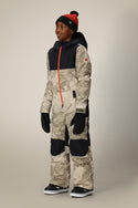 A young person wears the 686 Boys Shazam Insulated One-Piece—a camo-patterned polyester snowsuit with black panels, black hood and gloves, plus a black beanie with red stripe and pom-pom. The coverall features an orange front zipper.