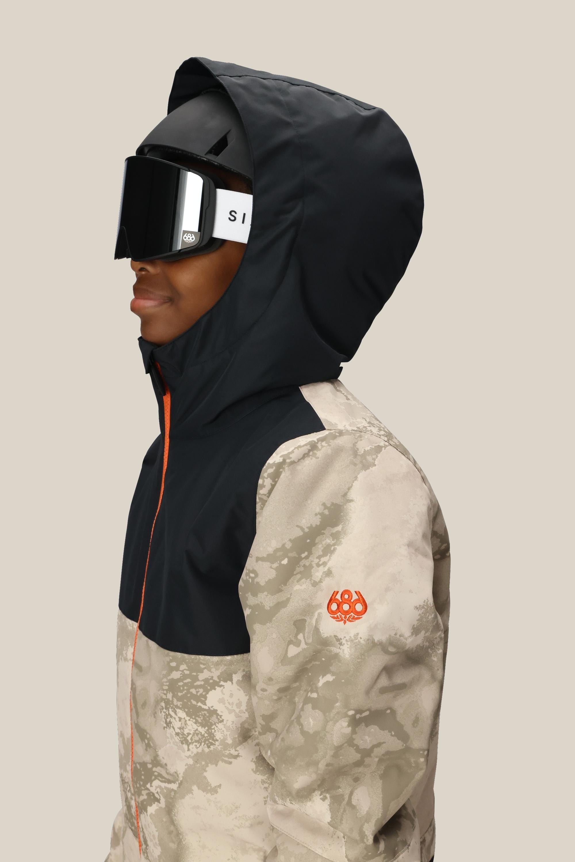 A person wears the 686 Boys Shazam Insulated One-Piece by 686, featuring a black hood, beige camo pattern, orange zipper, and large black ski goggles, standing against a plain light background.