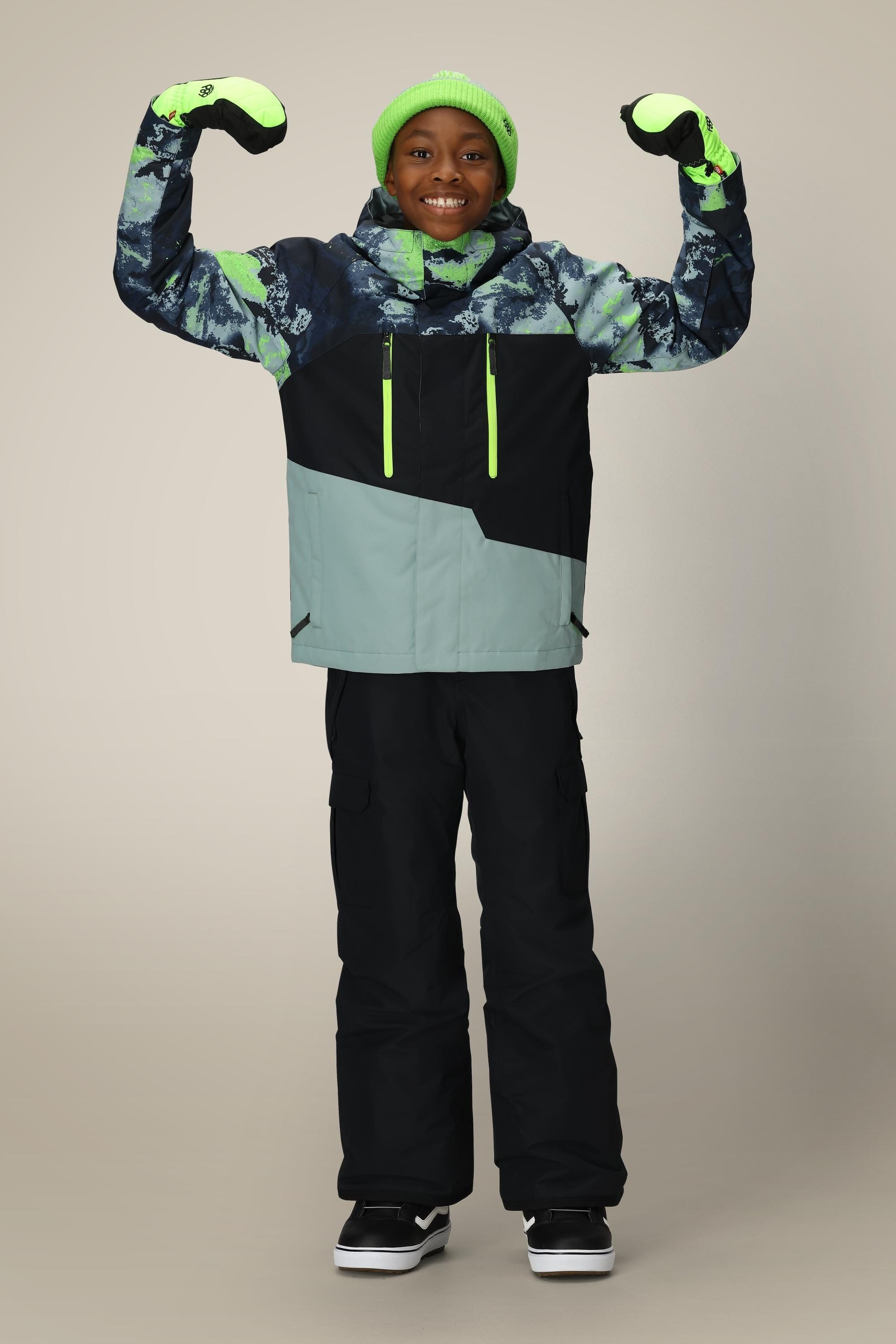 A smiling child wears the 686 Boys' Geo™ Insulated Jacket, black snow pants, neon green hat, and gloves, arms flexed confidently against a plain background—ready for cozy winter fun in this stylish outerwear from 686.