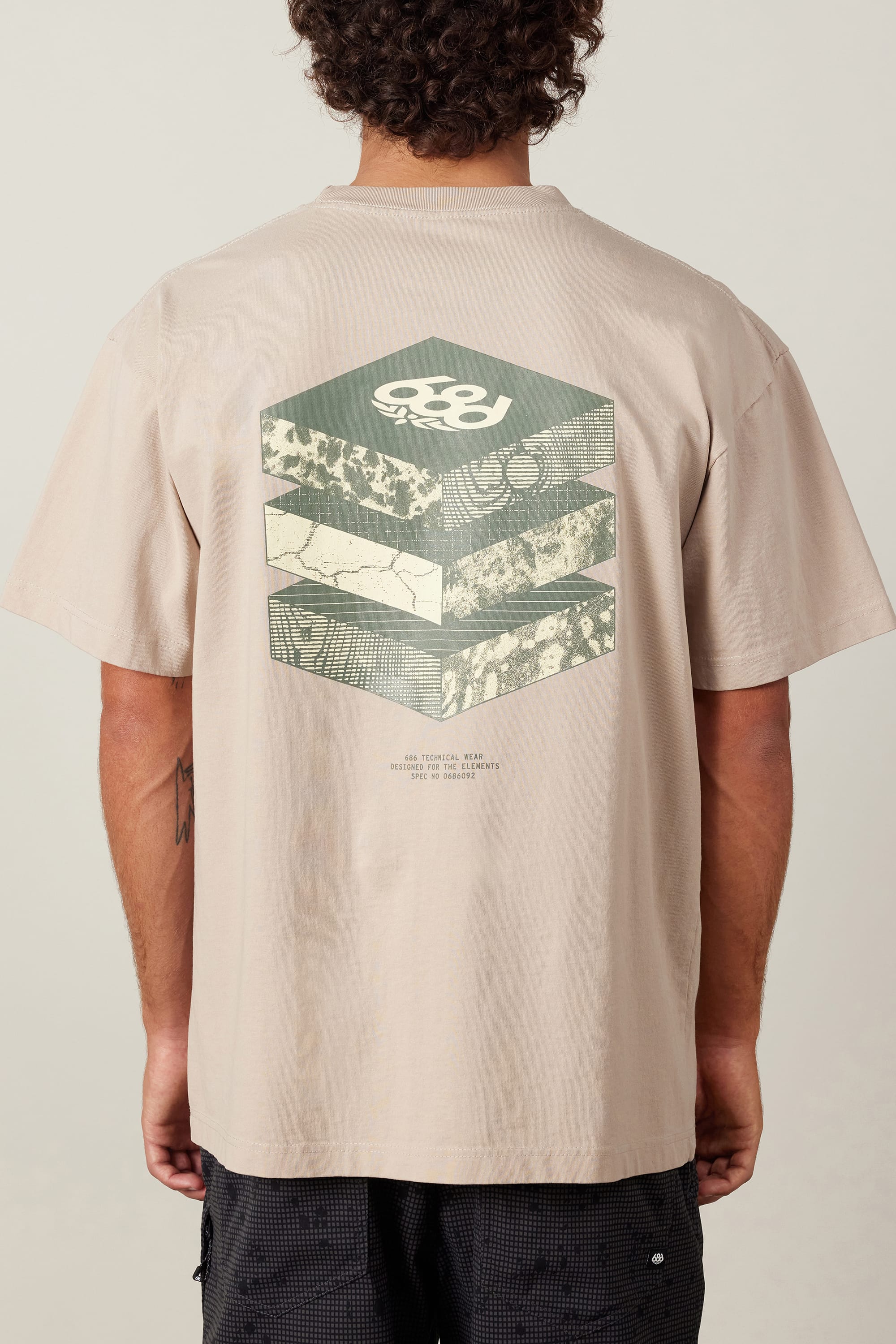 A person wears the 686 Elements Relaxed S/S Tee by 686, a beige men’s t-shirt made from 100% cotton, featuring geometric hexagonal graphics and layered abstract patterns with a small text section on the back.