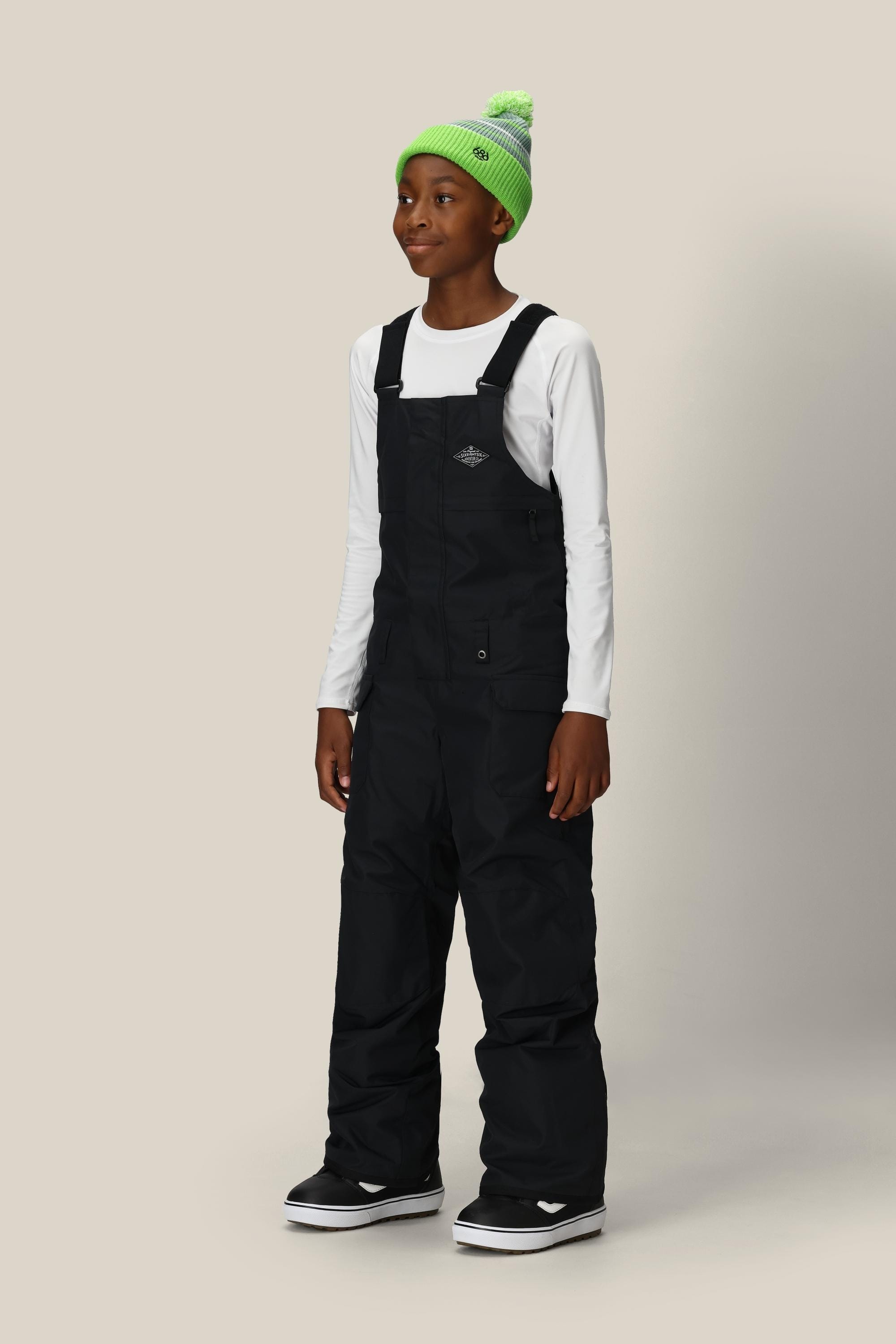 A smiling child wears the 686 Boys' Frontier™ Insulated Bib in black, styled with a bright green beanie and black boots, standing against a plain light background.