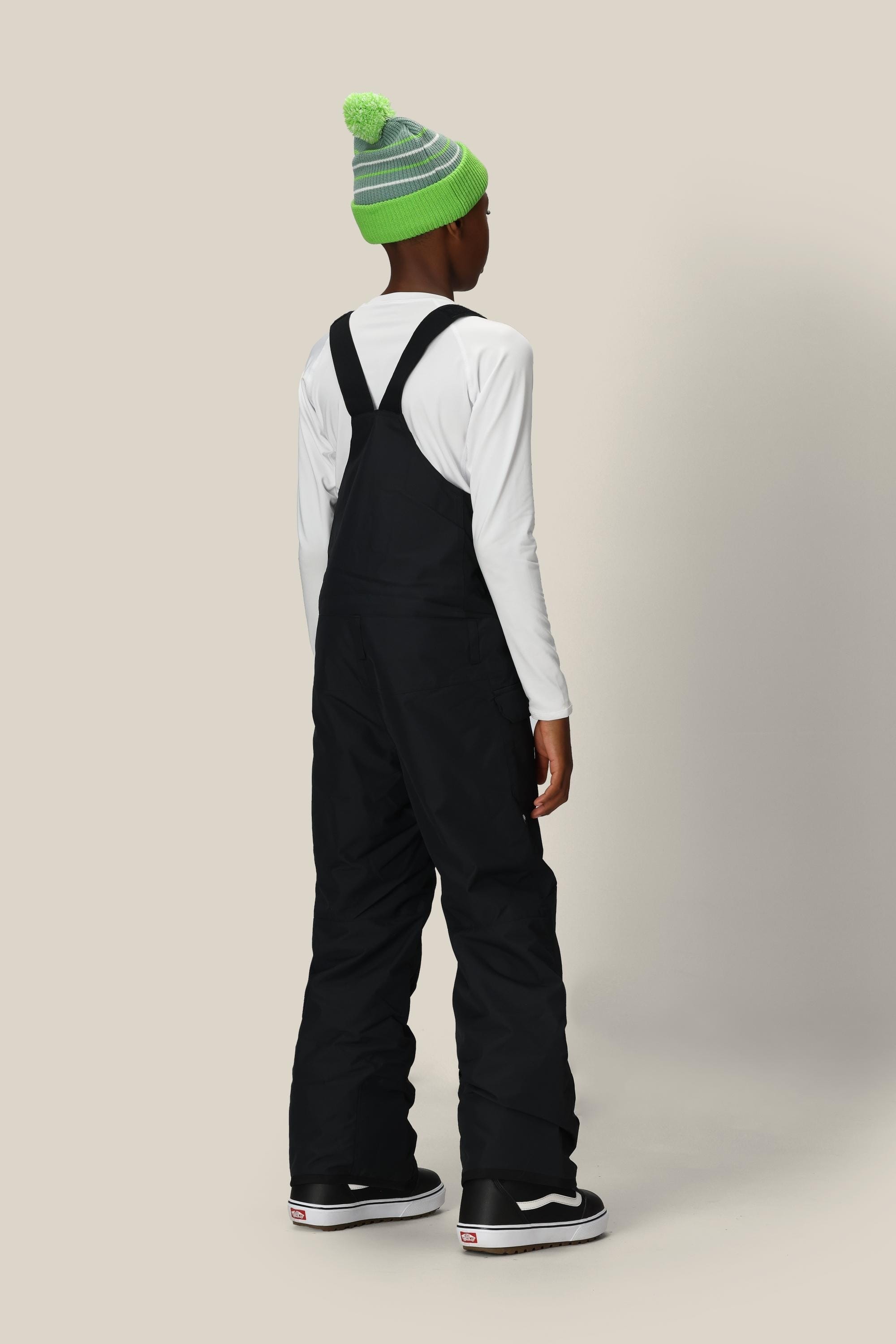 A child wearing the 686 Boys' Frontier™ Insulated Bib from 686 stands facing away, displaying the back of the black snow bib overalls, paired with a green and gray striped beanie and a white long-sleeve shirt against a beige background.