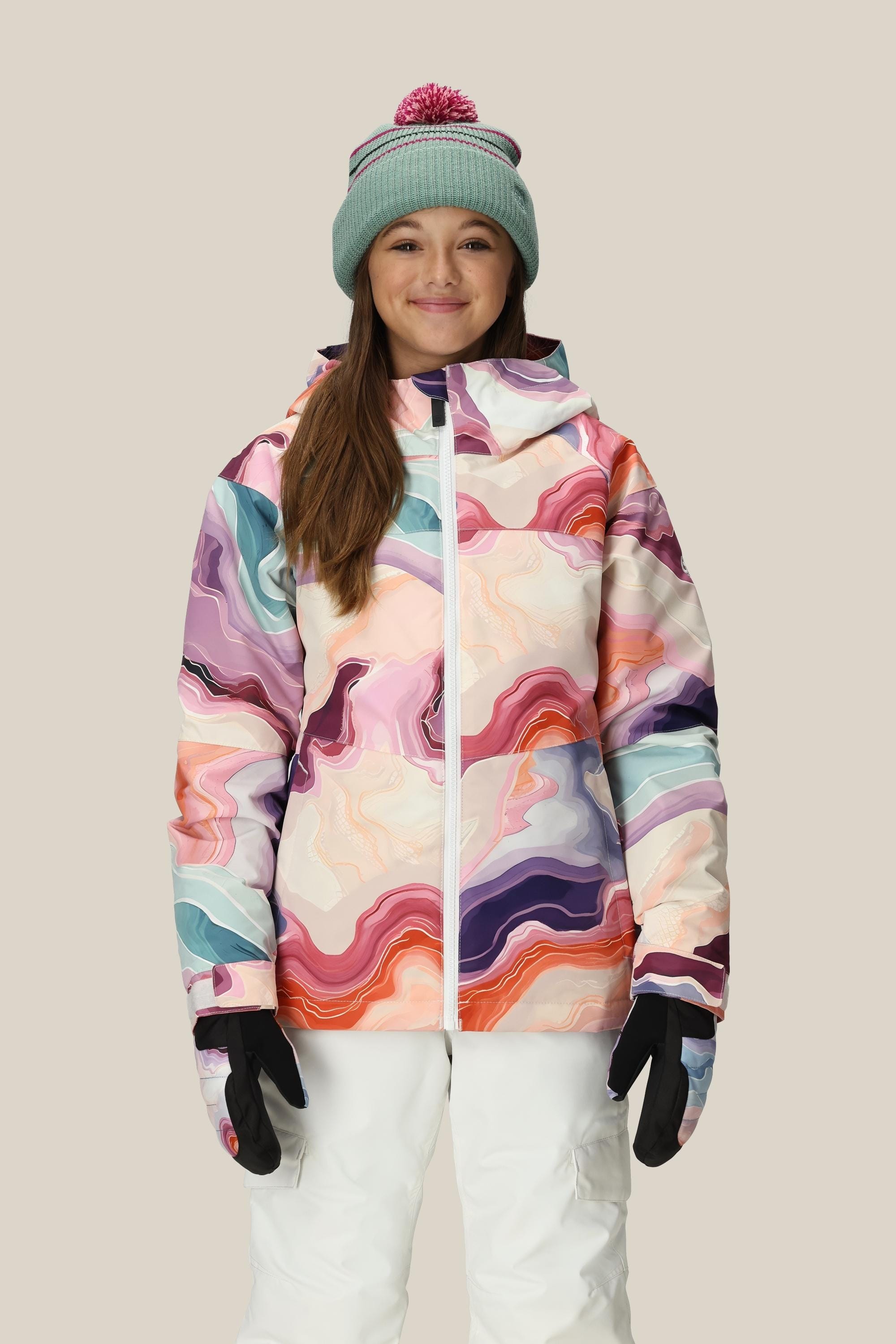 A person smiles, wearing the 686 Girls' Athena™ Insulated Jacket by 686 with colorful abstract patterns, a light blue beanie with a pink pom-pom, white snow pants, and black gloves against a plain beige background.