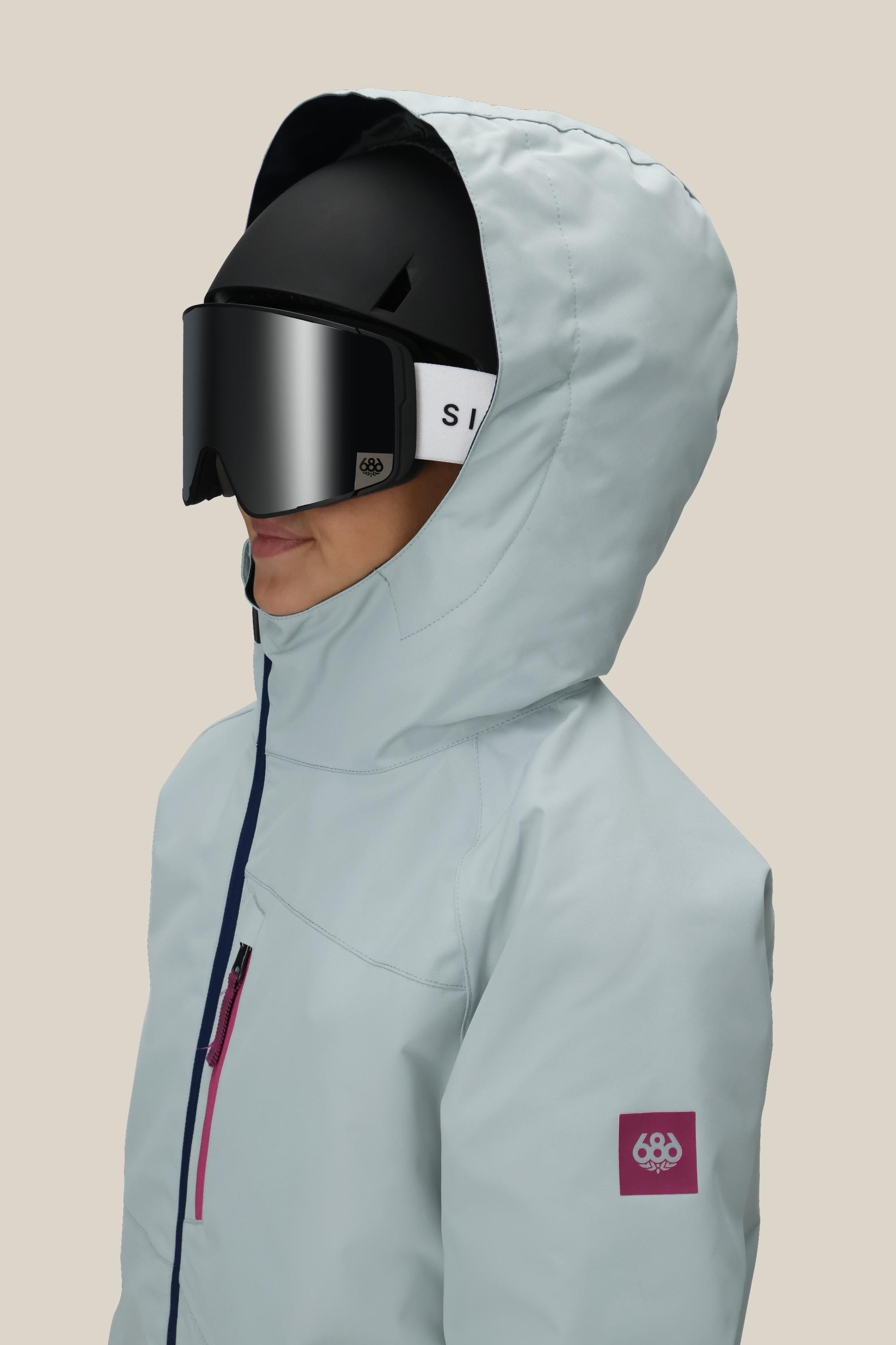 A girl wearing the 686 Girls' Hydra™ Insulated Jacket in light blue with body-mapped lining, chest zipper, and a small sleeve patch, paired with a black ski helmet and goggles. Side view against a beige background.