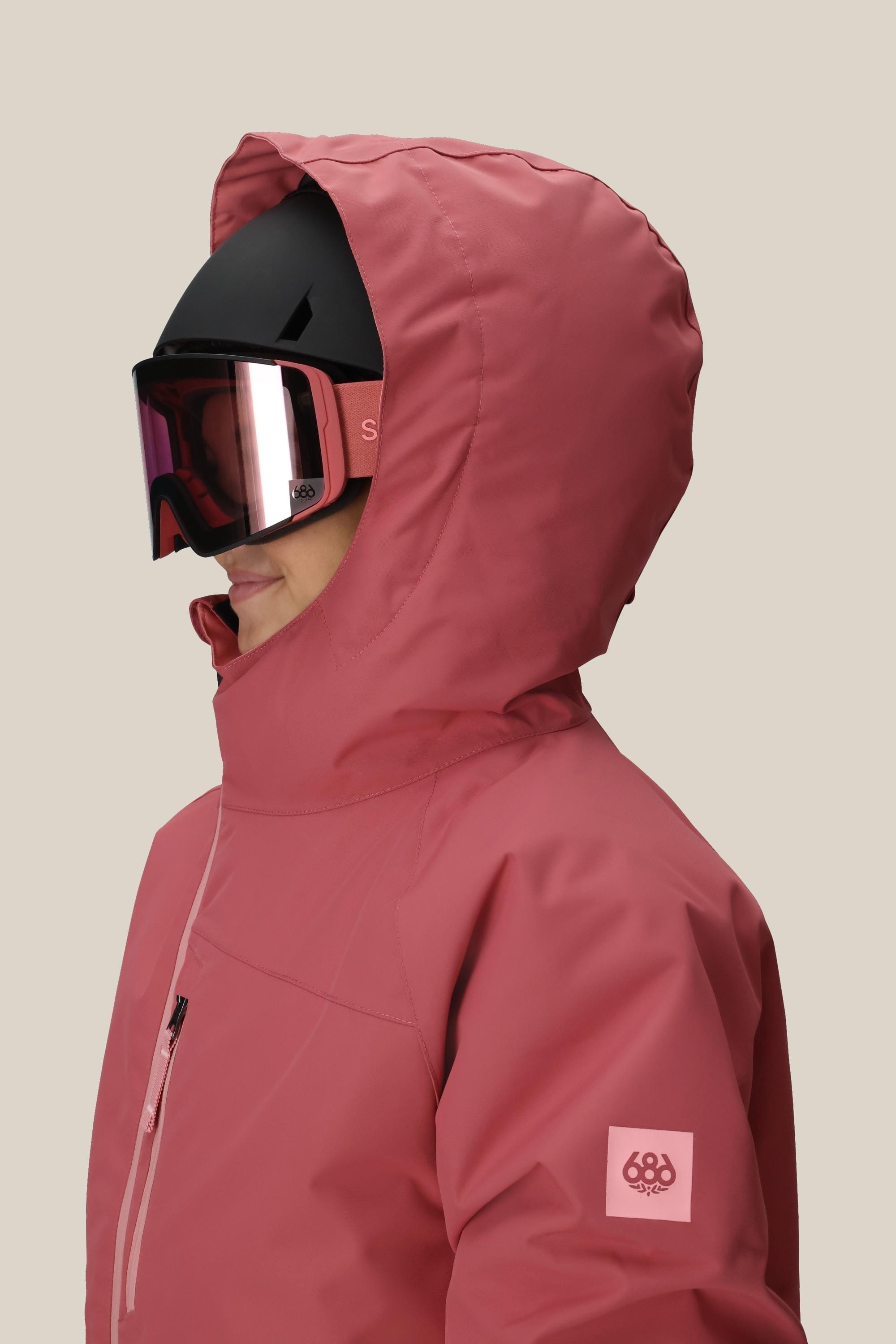 A girl wears the 686 Girls' Hydra™ Insulated Jacket in pink, featuring body-mapped lining for warmth, paired with a black ski helmet and reflective goggles, shown in profile against a plain beige background.