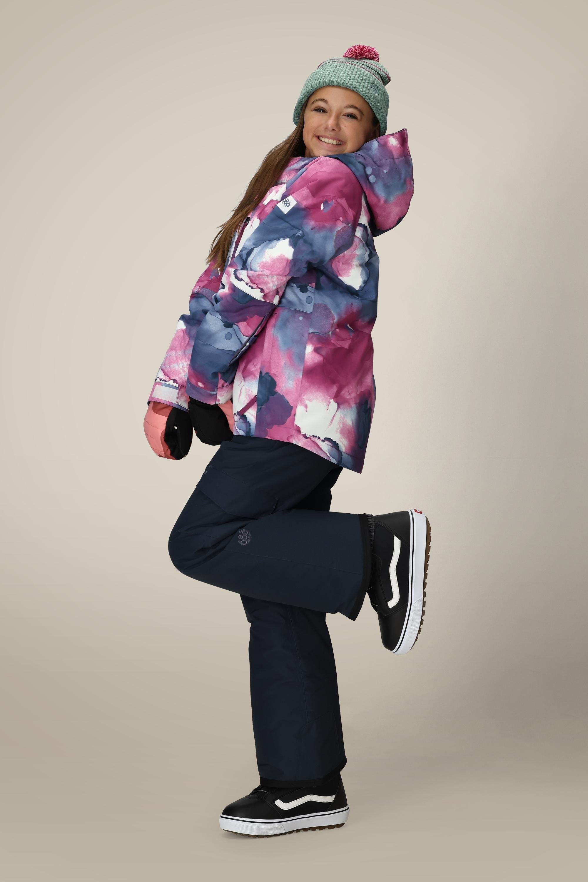 A smiling woman in a gray beanie playfully poses in the 686 Girls' Hydra™ Insulated Jacket by 686, featuring body-mapped lining for warmth. She completes her look with dark snow pants and black shoes against a plain background.