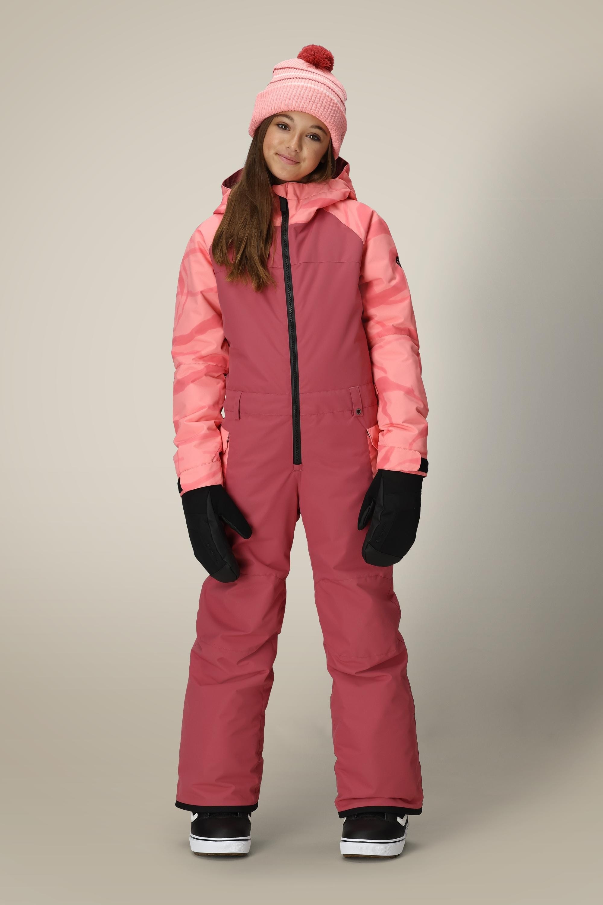 A young girl smiles, wearing the 686 Girls Shine Insulated One-Piece snowsuit in pink by 686, paired with black gloves and a pink knit pom-pom hat, standing against a plain light background.