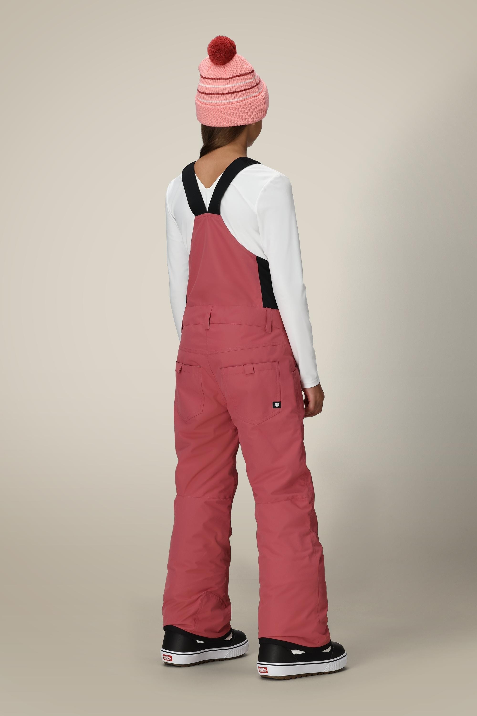 A child faces away, wearing a pink pom-pom beanie, white long-sleeve shirt, black and white sneakers, and 686 Girls Sierra Insulated Bib by 686 with black straps, shown against a plain background.