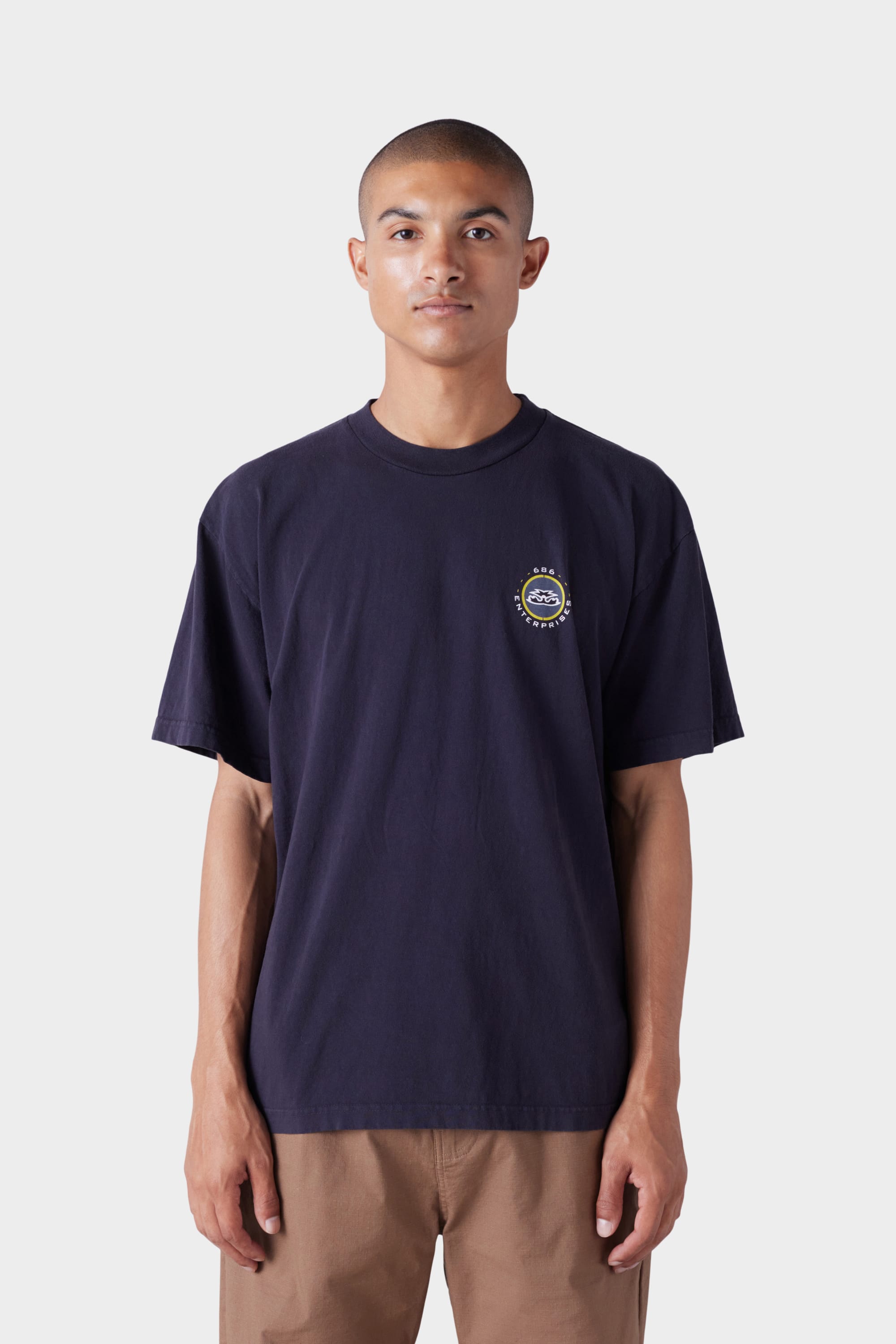 A person stands against a plain light background wearing the 686 Mens Jib 2001 Badge Short Sleeve Tee by 686 in navy blue with a small circular left-chest logo, paired with light brown pants.
