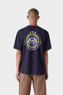 A person with short hair is seen from behind wearing the 686 Mens Jib 2001 Badge Short Sleeve Tee in dark blue, featuring a large circular 686 logo and graphic on the back, paired with brown pants.