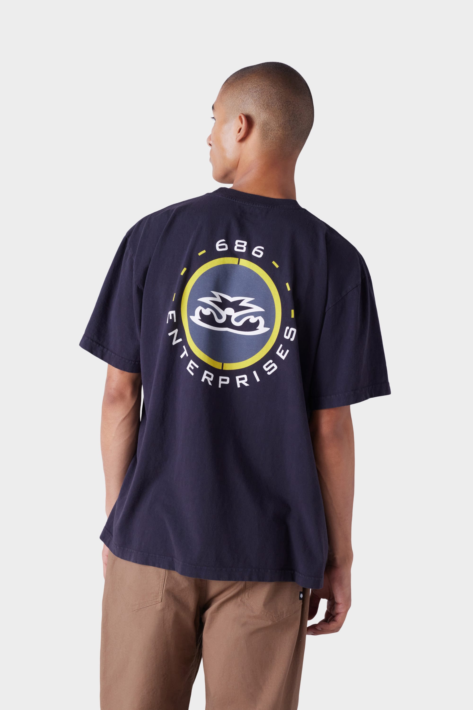 A man is seen from behind wearing the 686 Mens Jib 2001 Badge Short Sleeve Tee in navy blue, featuring a large circular 686 ENTERPRISES logo on the back, paired with brown pants.