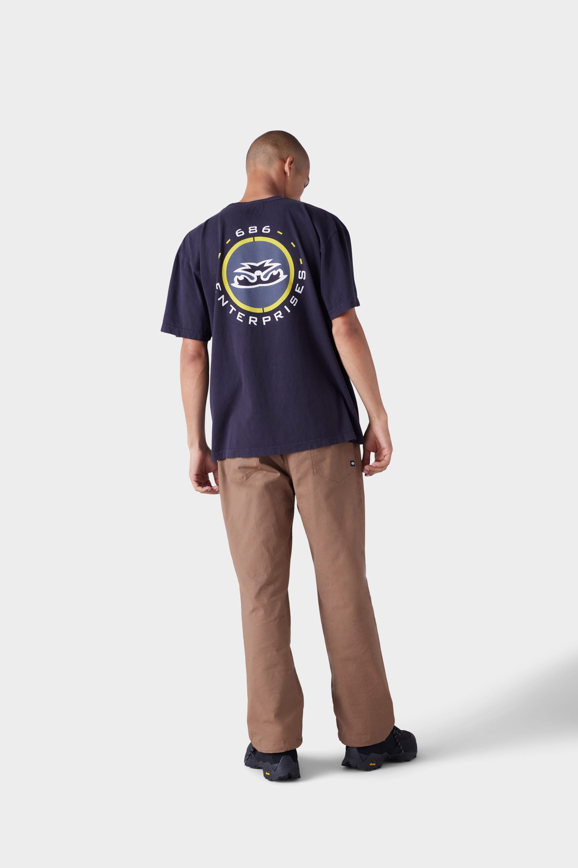 A person stands facing away, wearing the 686 Mens Jib 2001 Badge Short Sleeve Tee in navy with a circular yellow and white back graphic, brown pants, and black shoes, against a plain light gray background.