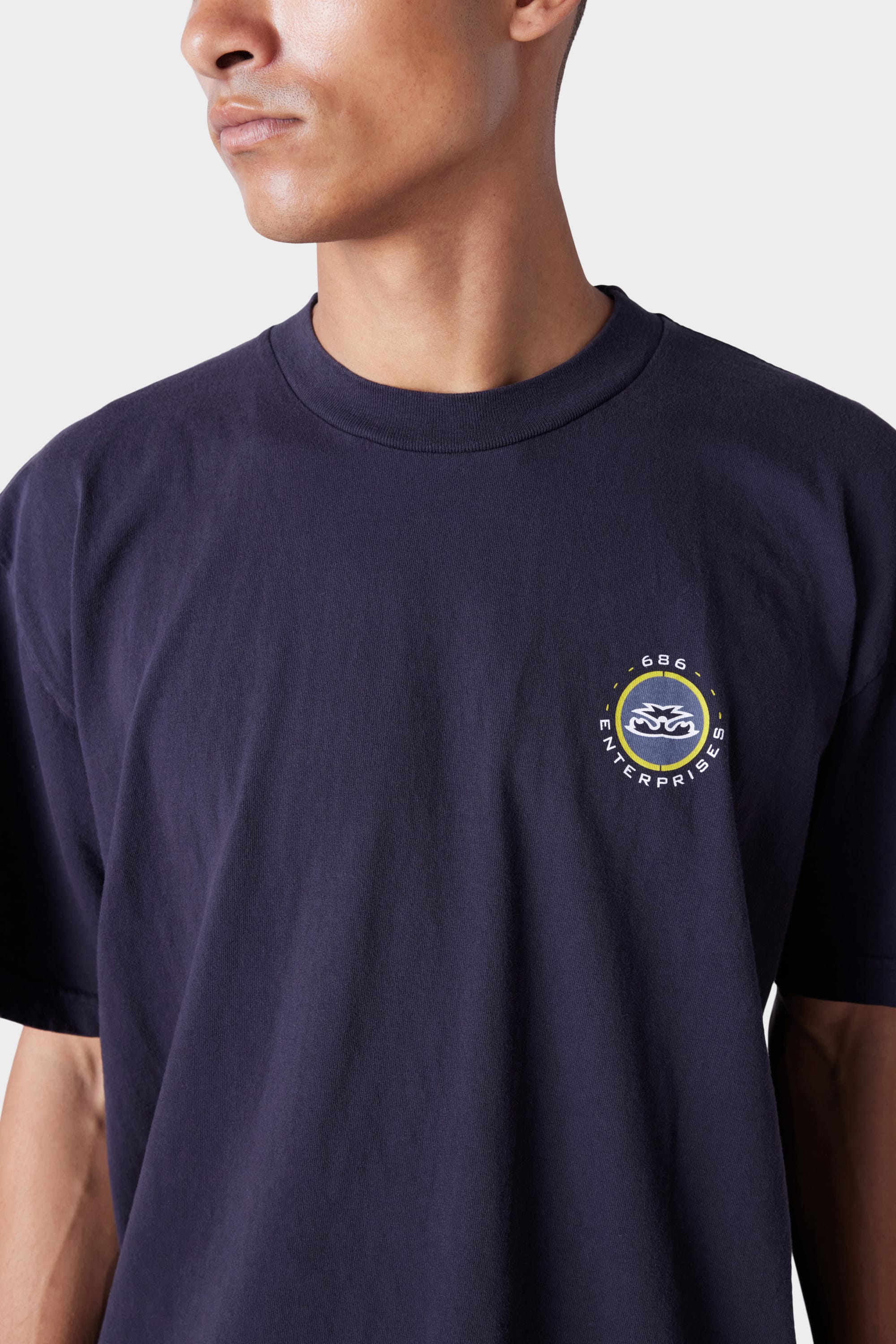 A person wears the 686 Mens Jib 2001 Badge Short Sleeve Tee in dark navy, featuring a small left-chest circular 686 Enterprises logo with stylized waves. They are facing slightly to the side. Brand: 686.