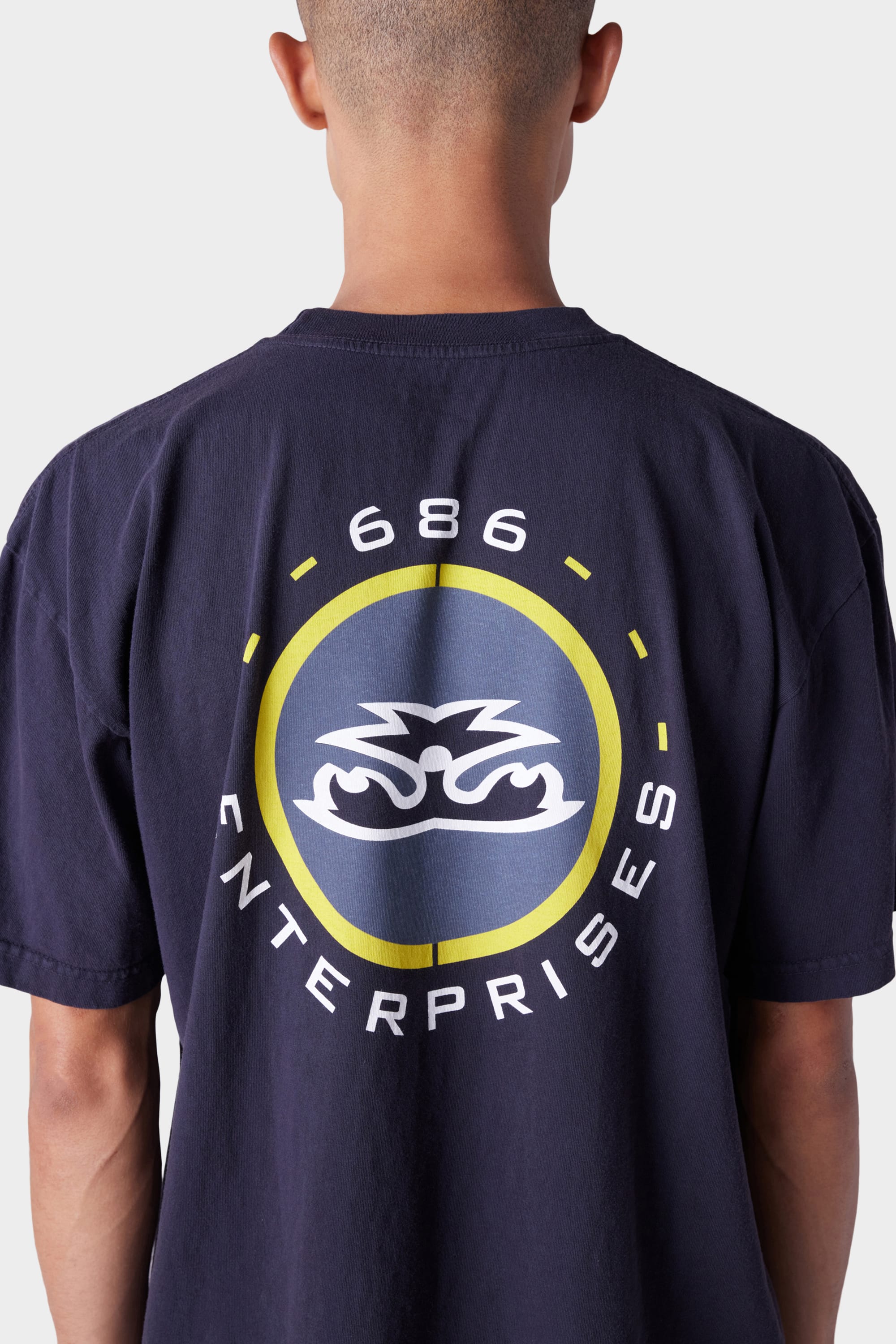 A person seen from the back wearing the 686 Mens Jib 2001 Badge Short Sleeve Tee in navy blue, featuring a round yellow and blue logo with 686 ENTERPRISES text and a stylized design.