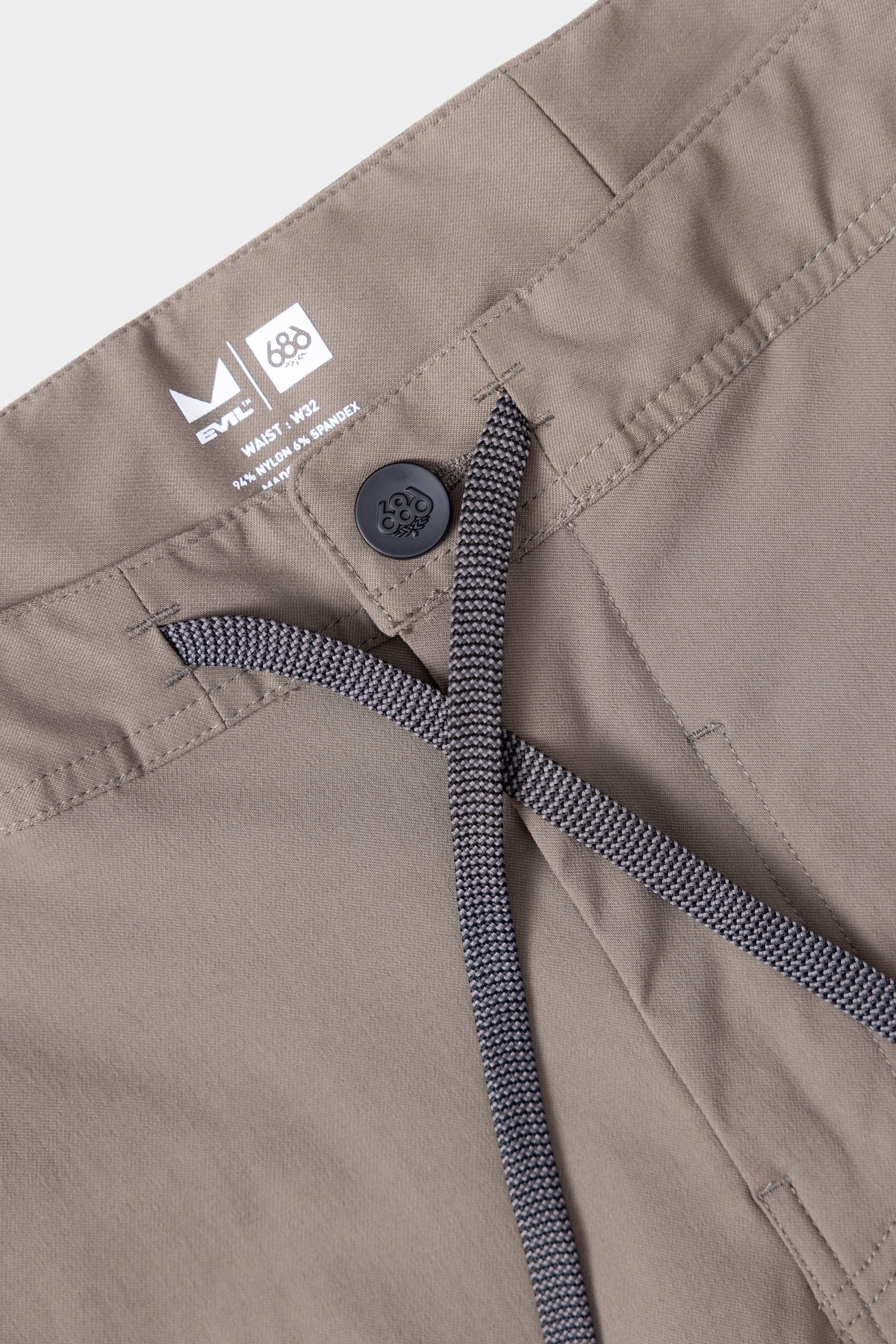 Close-up of 686 Mens Platform Bike Pant - Relaxed Fit waistband shows black button, gray drawstring, and inner tag with 686 branding and size. Durable fabric with detailed stitching around the waistband is visible.