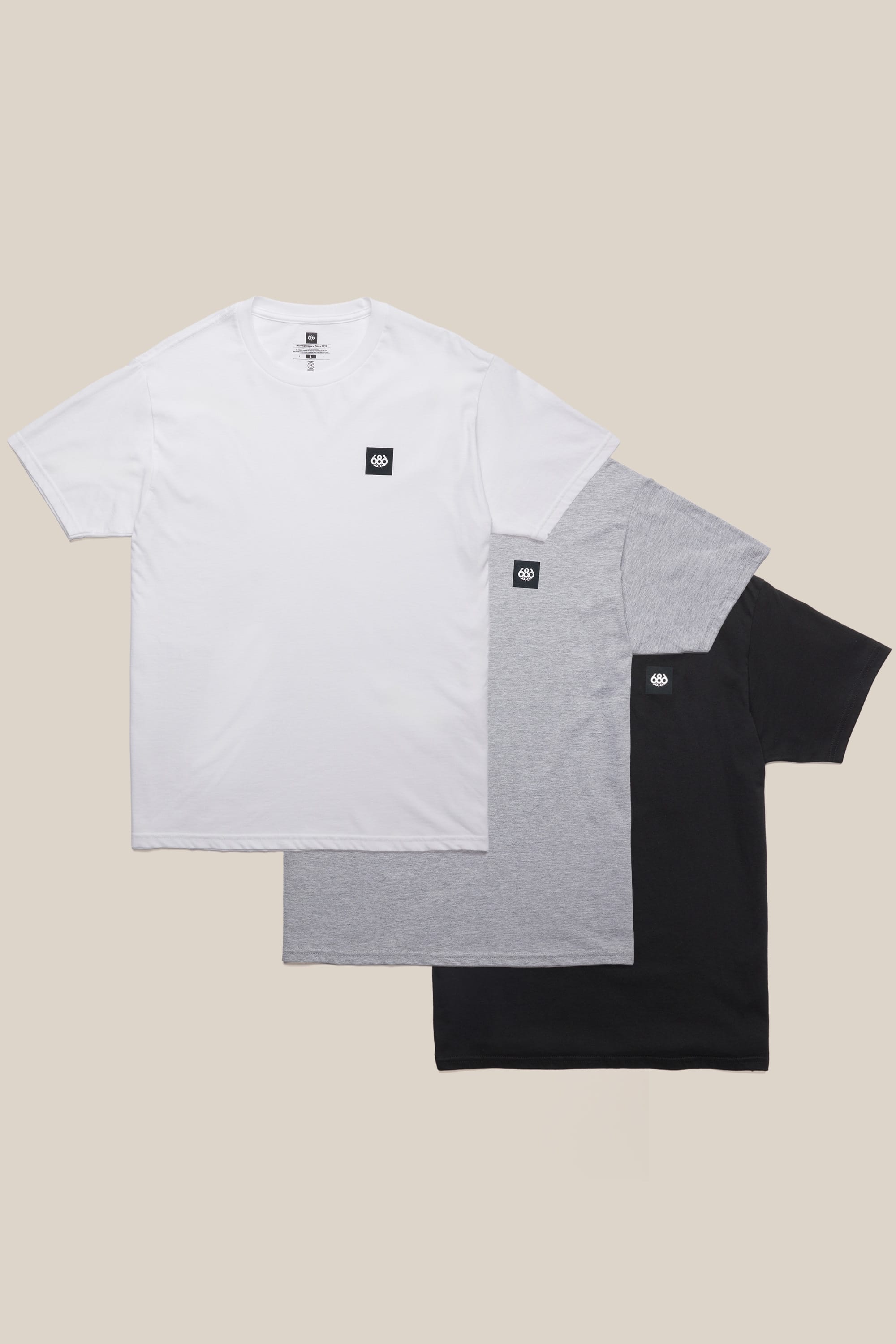 Three men's crew-neck tees from the 686 Logo 3-Pack S/S Tee set in white, gray, and black overlap on a beige background. Each 100% cotton tee by 686 features a small black patch with a white logo on the left chest.