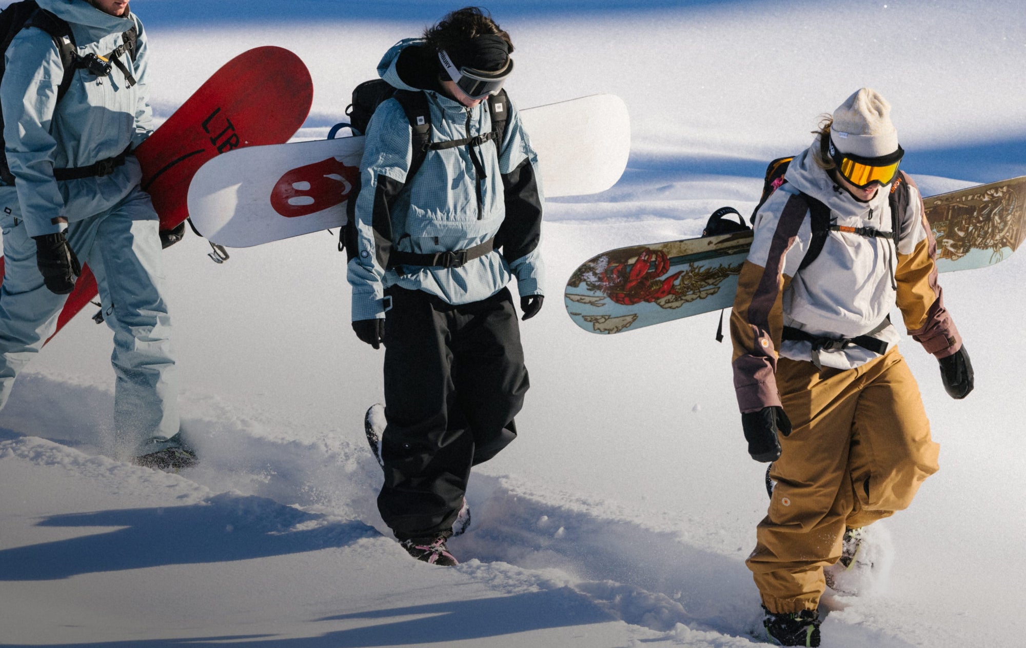 Three people in winter gear walk through snow, each carrying a snowboard on their back. The sun casts shadows, and the scene suggests they are headed to a snowboarding spot.