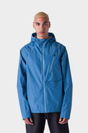A person with short blond hair wears the 686 Men's Hydra™ 2.5L All-Weather Jacket by 686, featuring a blue hood and zippered chest pocket, paired with black pants, standing against a plain white background.