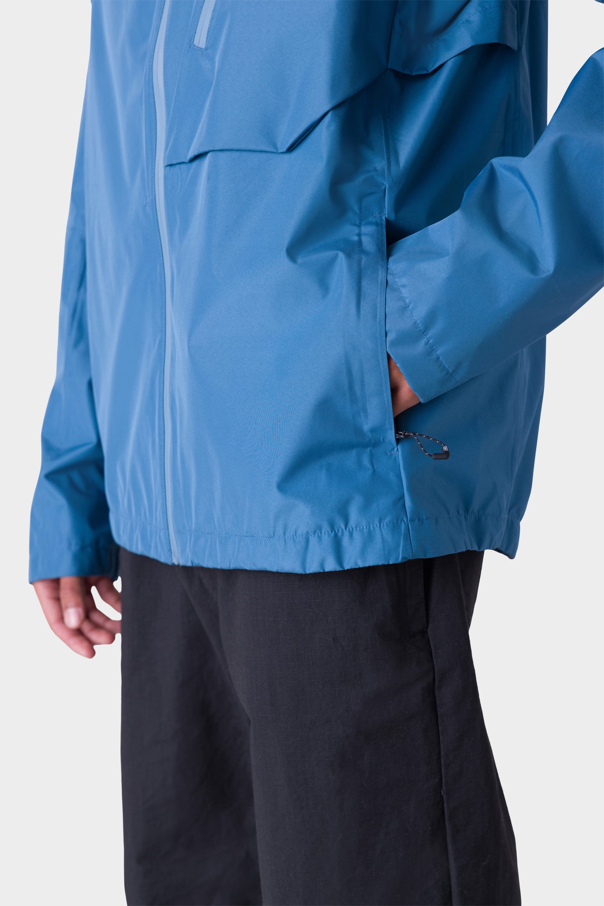 A person wearing the 686 Men's Hydra™ 2.5L All-Weather Jacket in blue, hands in side pocket and paired with black pants; image is cropped to show only upper legs and torso against a plain light background.