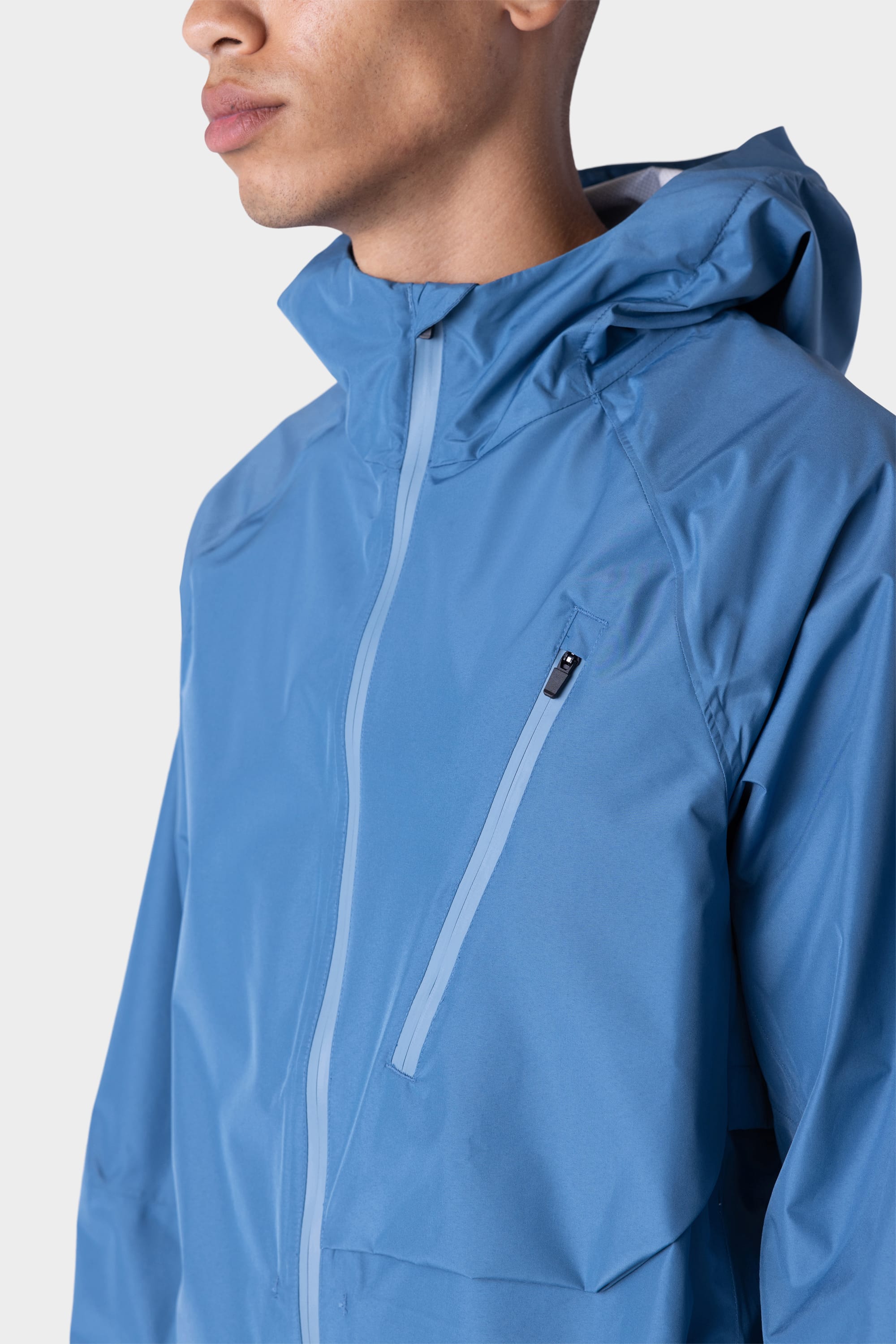 A person wears the 686 Men's Hydra™ 2.5L All-Weather Jacket—a blue hooded rain jacket with a zippered chest pocket—shown from the shoulders up against a plain light background.