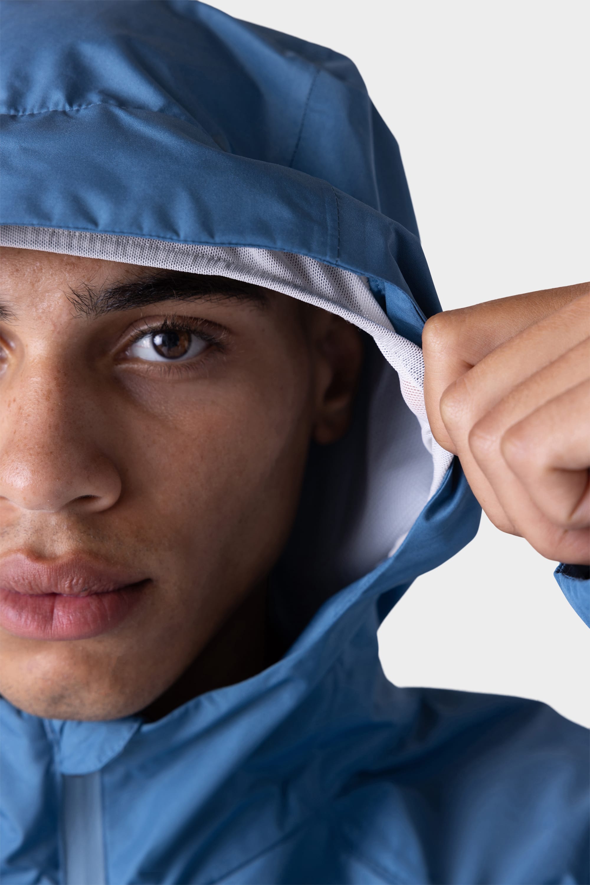 Close-up of a person wearing the 686 Men's Hydra™ 2.5L All-Weather Jacket in blue, pulling the edge of the hood with one hand against a plain light background, with only half of their face visible.