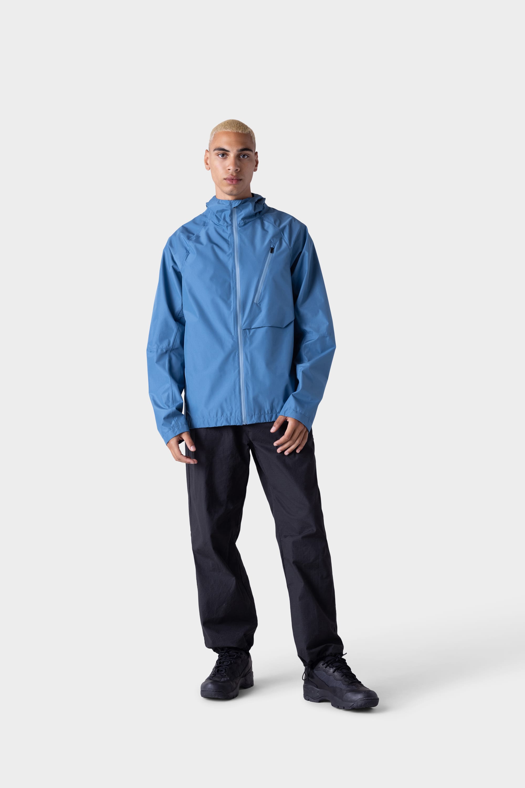 A person with short blond hair wears the 686 Men's Hydra™ 2.5L All-Weather Jacket by 686 in light blue, paired with black pants and black shoes, standing against a plain light gray background.
