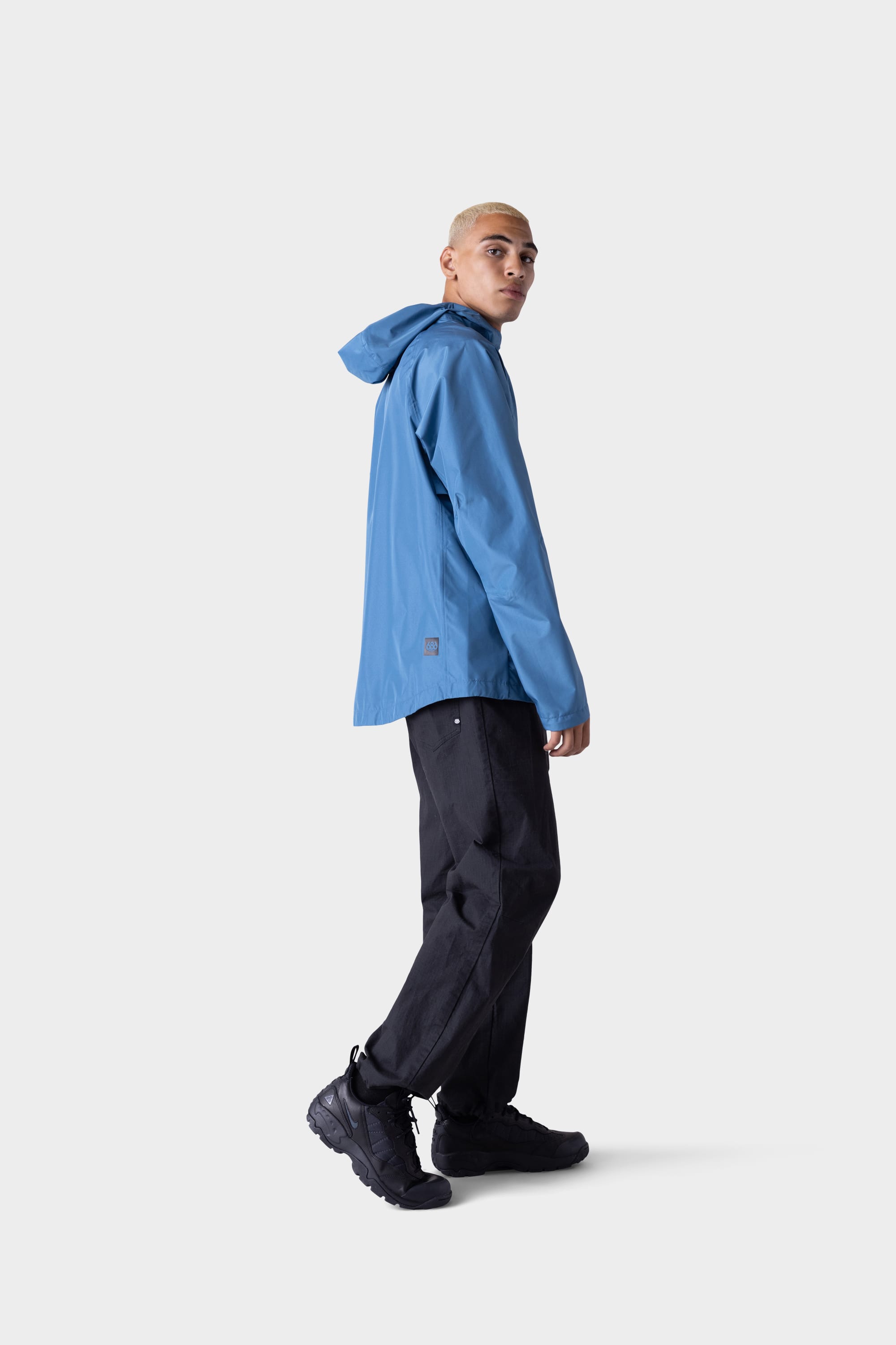 A person with short blond hair models the 686 Men's Hydra™ 2.5L All-Weather Jacket in blue, paired with black pants and sneakers, standing against a light gray background and glancing over their shoulder.