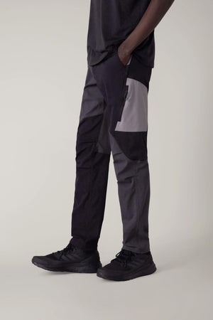 A person models the 686 Men's Anything Cargo Pant - Slim Fit in color-blocked black and gray, paired with black sneakers, hands in pockets, against a plain light background. Photo shows the pants from chest down, highlighting the 13-pocket design.