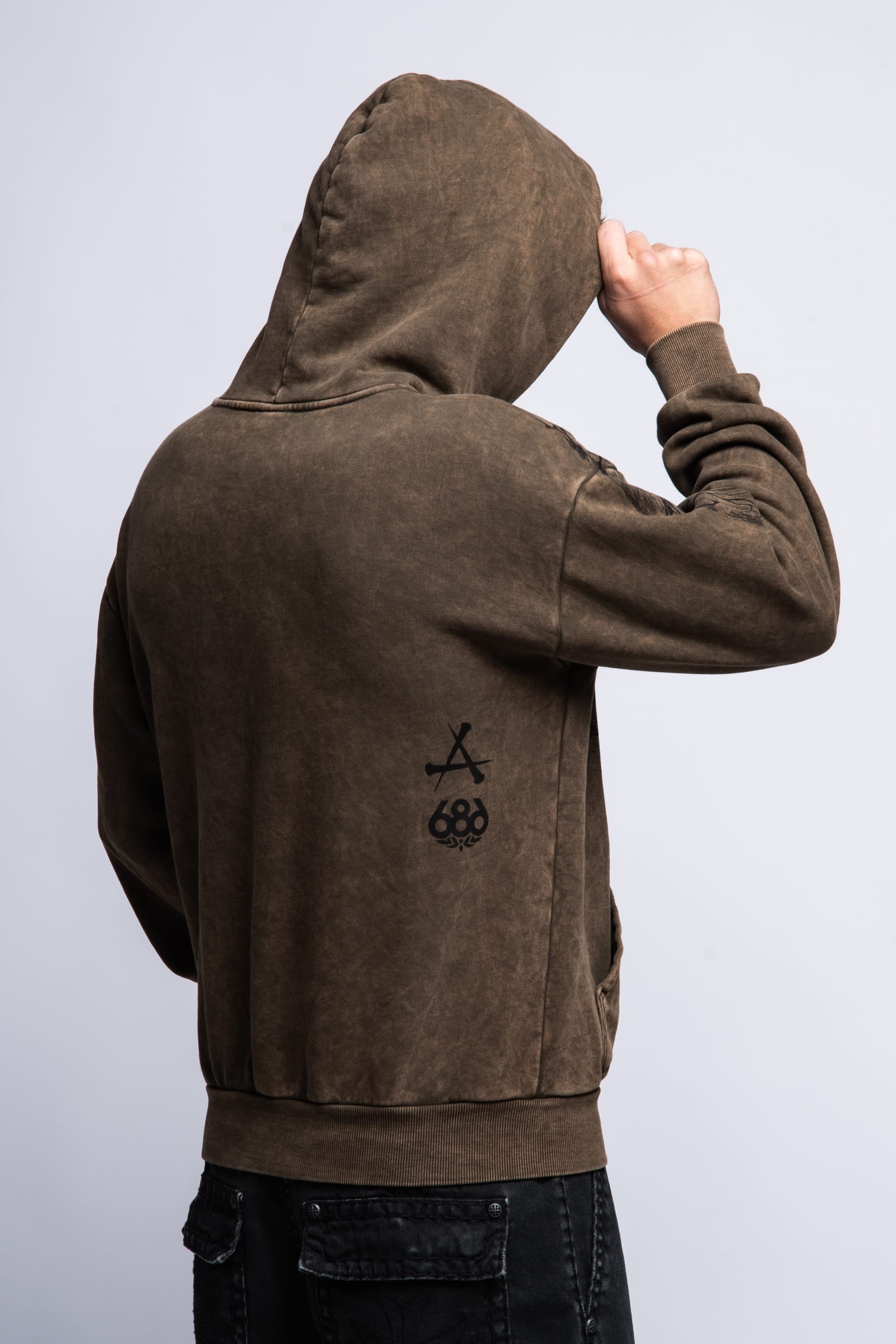 A person faces away from the camera wearing a 686 Affliction Natural Rot Pullover Hoody with the hood up, showing black graphics on the back and holding the hood’s edge with one hand.