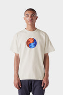 A person wearing the 686 Mens Bark and Bite Short Sleeve Tee by 686, featuring a cream color and a chest graphic of two stylized hands—one orange, one blue—reaching toward each other, stands against a plain white background.