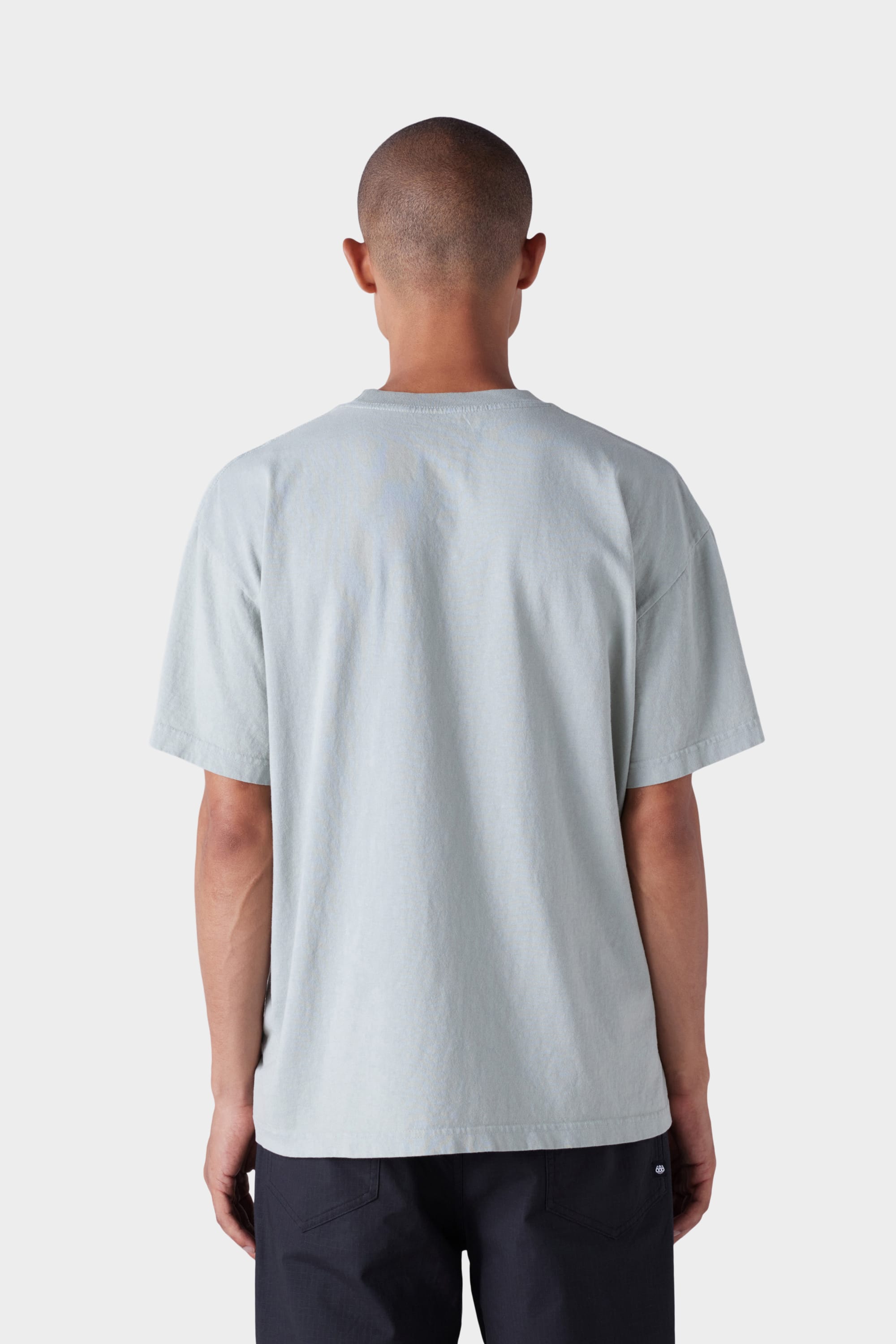 A person with a shaved head is seen from behind wearing the 686 Mens Blur Short Sleeve Tee in light gray and black pants, standing against a plain white background.