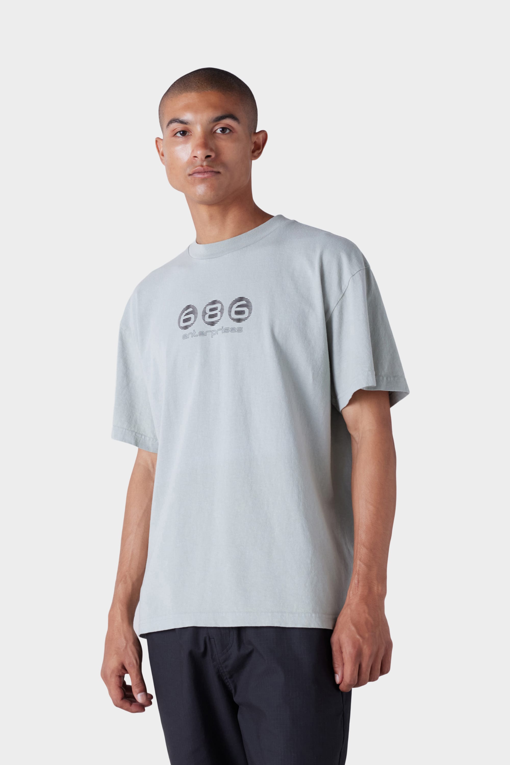 A person wearing the 686 Mens Blur Short Sleeve Tee by 686—an oversized light gray t-shirt with three black circular graphics and numbers on the chest—paired with black pants, stands against a plain light gray background.