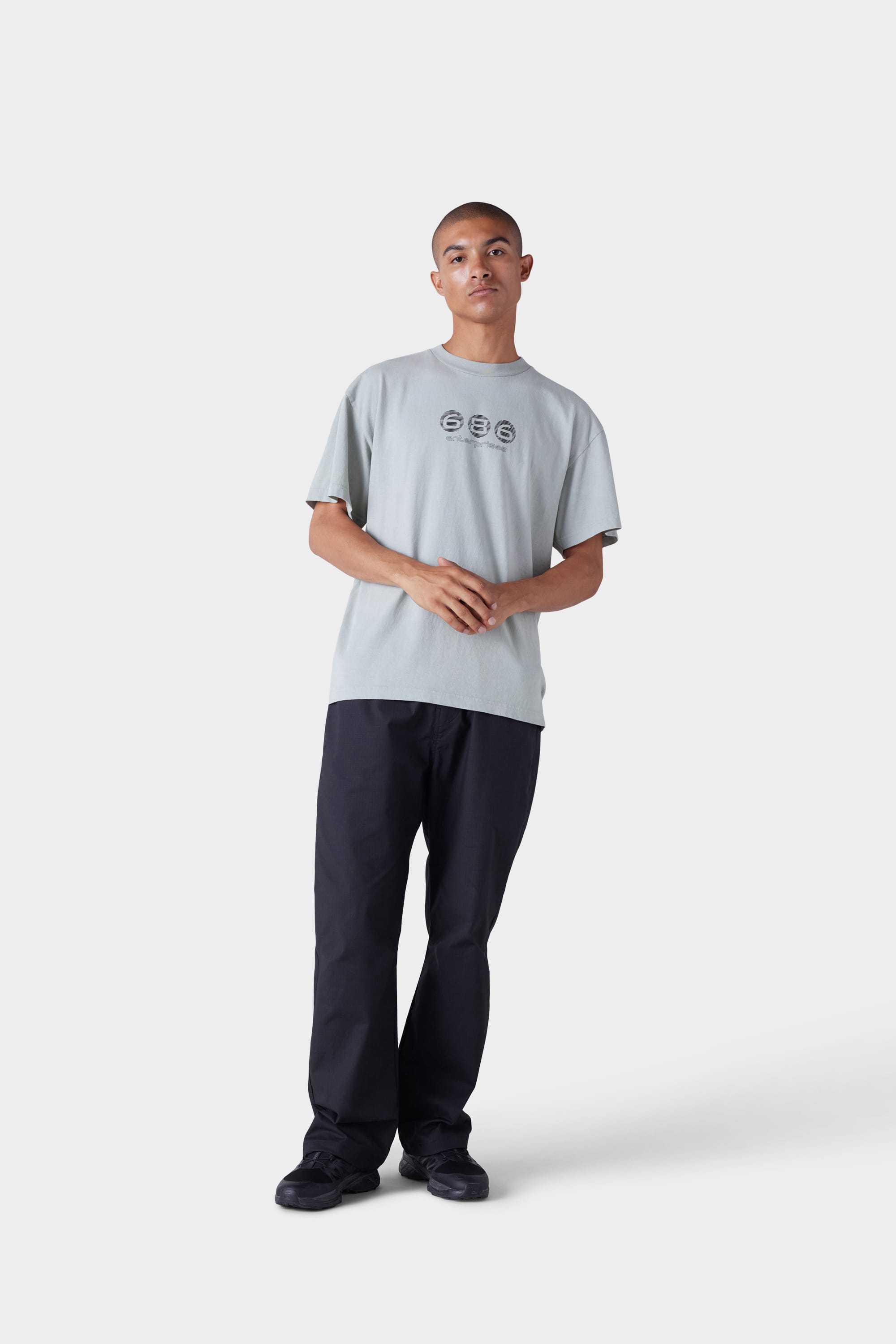 A person stands against a plain light grey background, wearing the 686 Mens Blur Short Sleeve Tee by 686, which features three circular designs on the chest, along with dark pants and black shoes. Their hands are gently clasped in front.