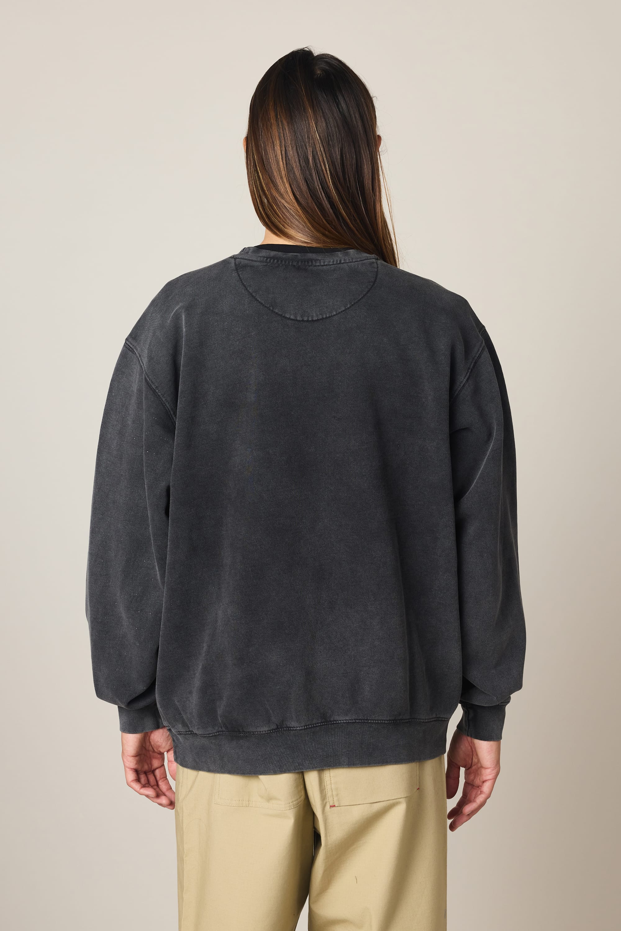 A person with long brown hair stands with their back to the camera against a plain light background, wearing the 686 Men's Mixed Relaxed LS Crewneck by 686 in faded black and beige pants.