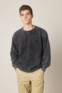 A young man with short curly hair wears the 686 Wreath Relaxed LS Crewneck by 686, a faded black 100% cotton sweatshirt, paired with beige pants, standing with his hands in his pockets against a plain light background.