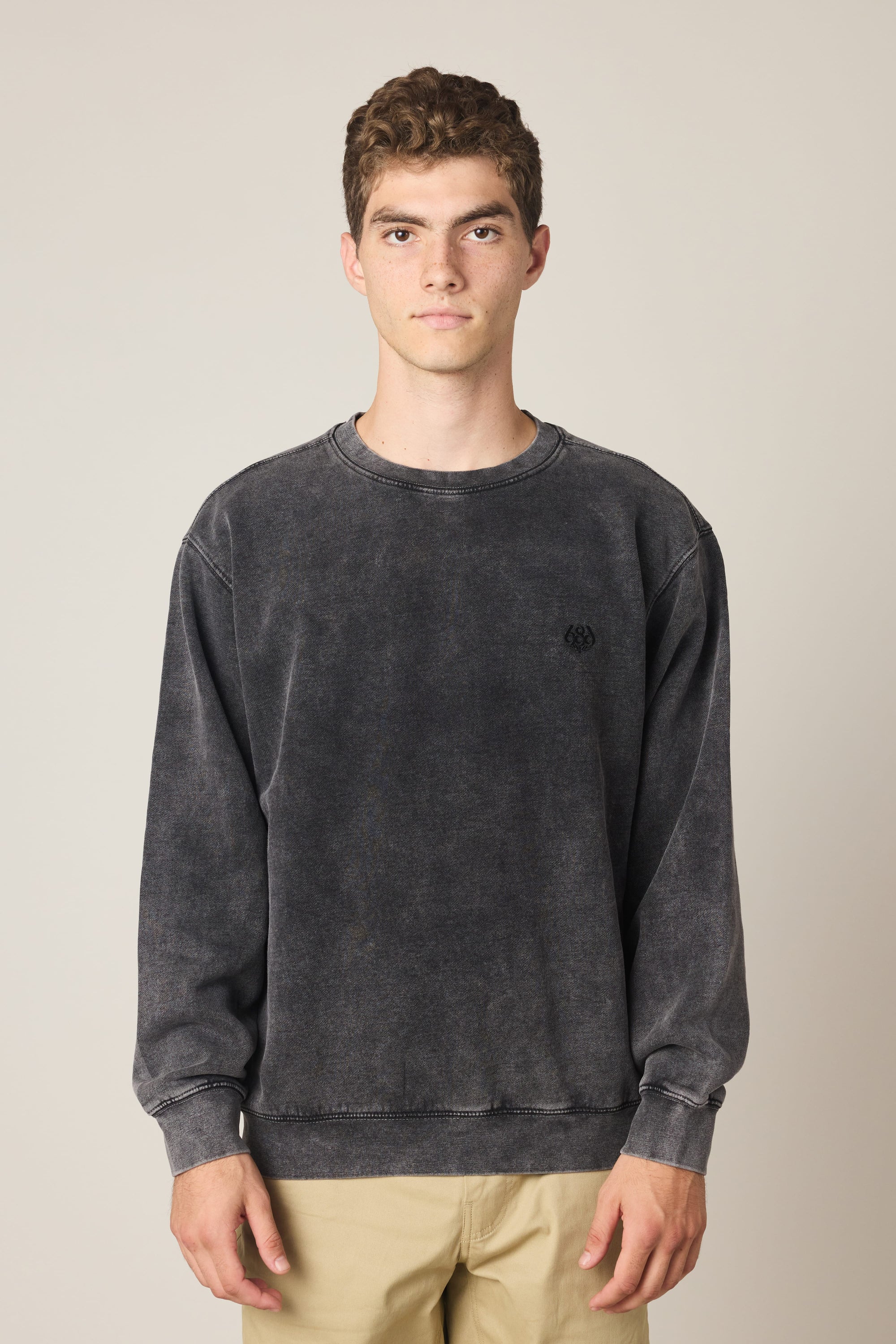 A young man with short curly hair wears the 686 Wreath Relaxed LS Crewneck sweatshirt in faded black and beige pants, standing against a plain light background and looking directly at the camera with a neutral expression.