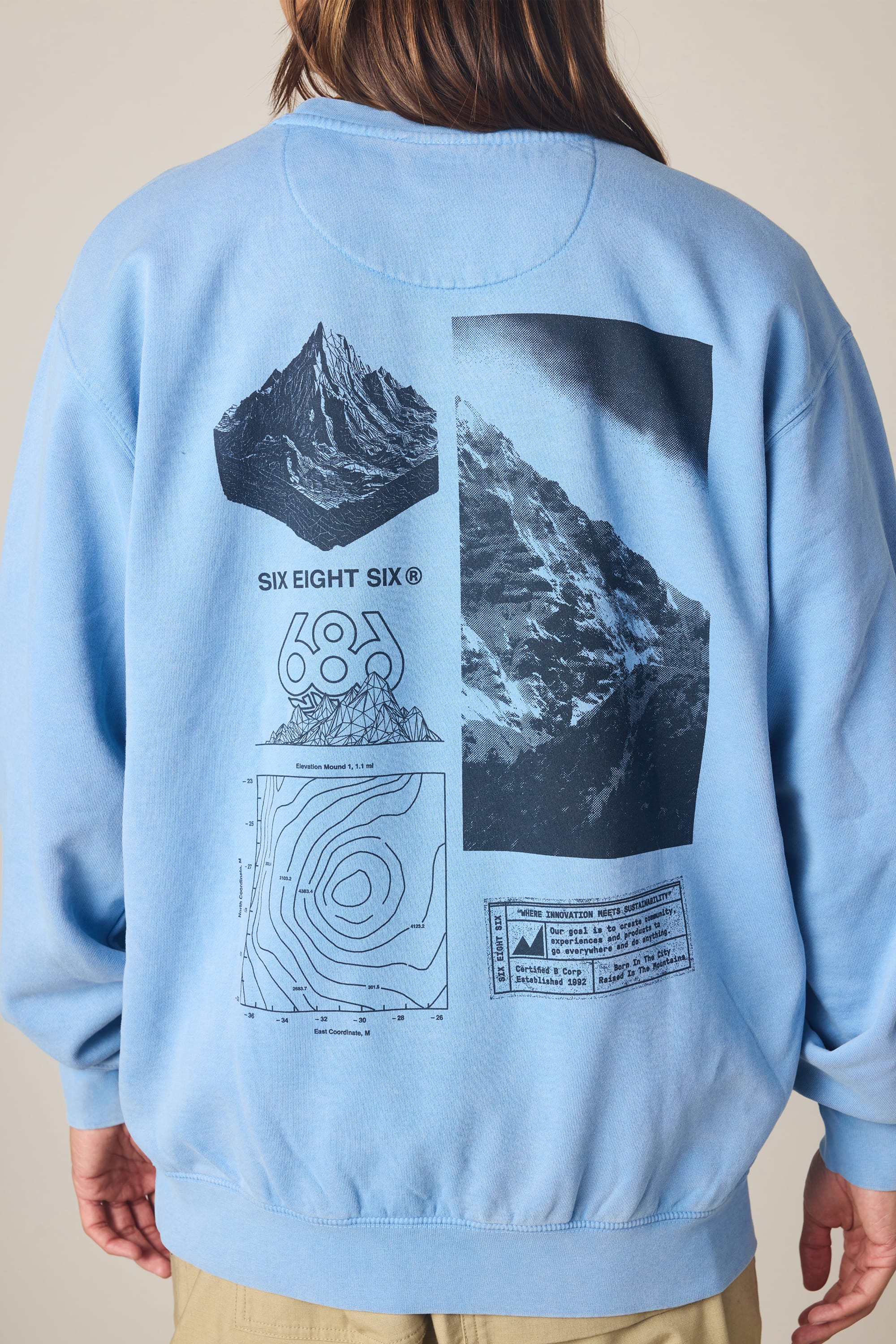 A person faces away from the camera, wearing a 686 Men's Mixed Relaxed LS Crewneck in light blue 100% cotton, featuring black mountain graphics, contour lines, and text on the back.