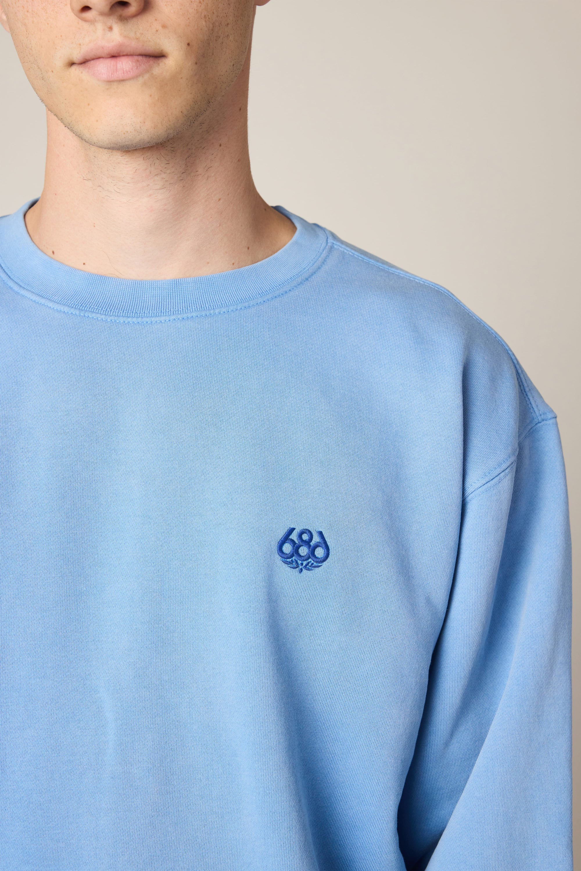 A person wears the 686 Wreath Relaxed LS Crewneck in light blue, a 100% cotton sweatshirt with a small dark blue embroidered 686 logo on the left chest, shown from the shoulders up against a neutral background.