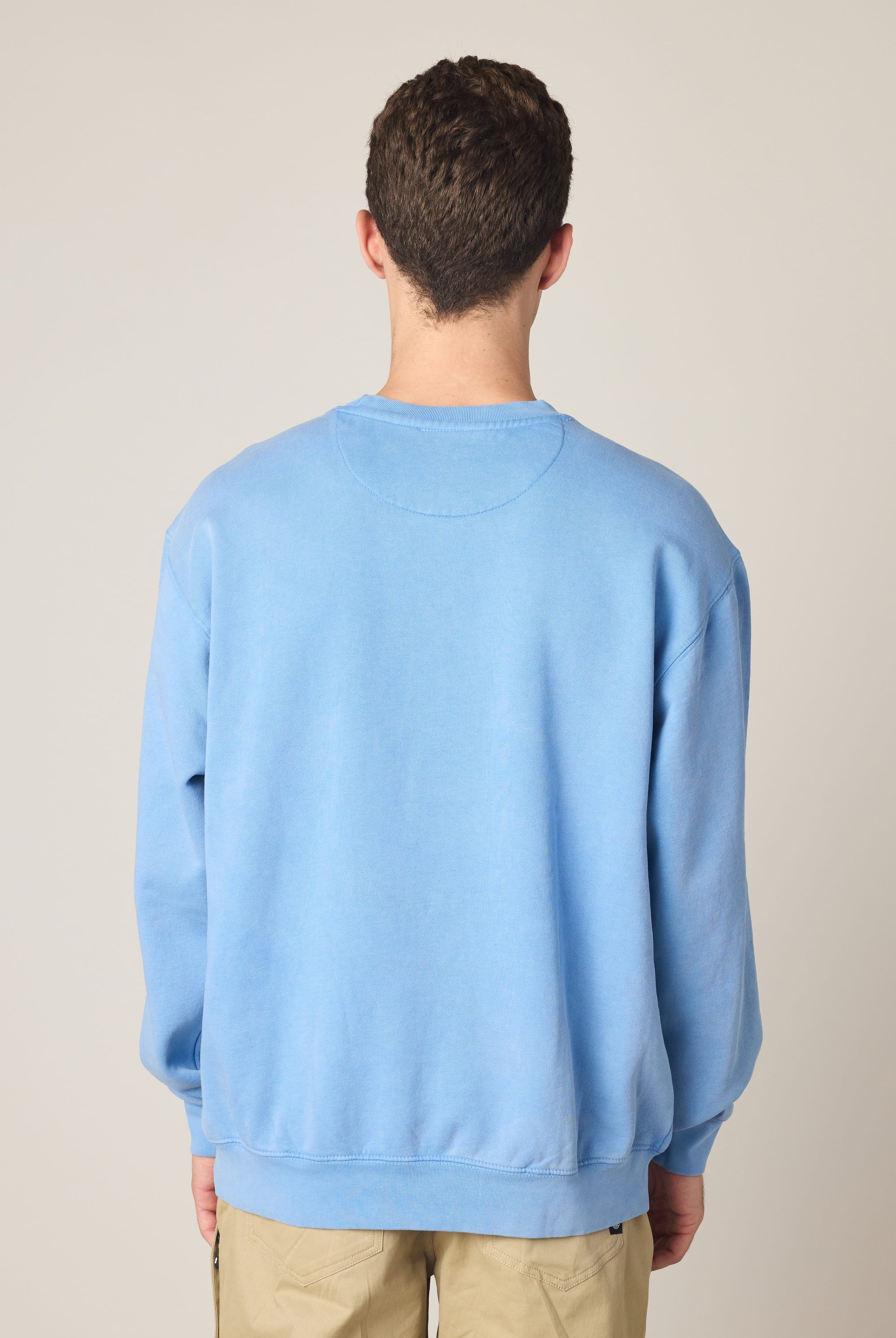 A person with short curly brown hair stands facing away from the camera, wearing the 686 Wreath Relaxed LS Crewneck by 686 and beige pants against a plain light background.