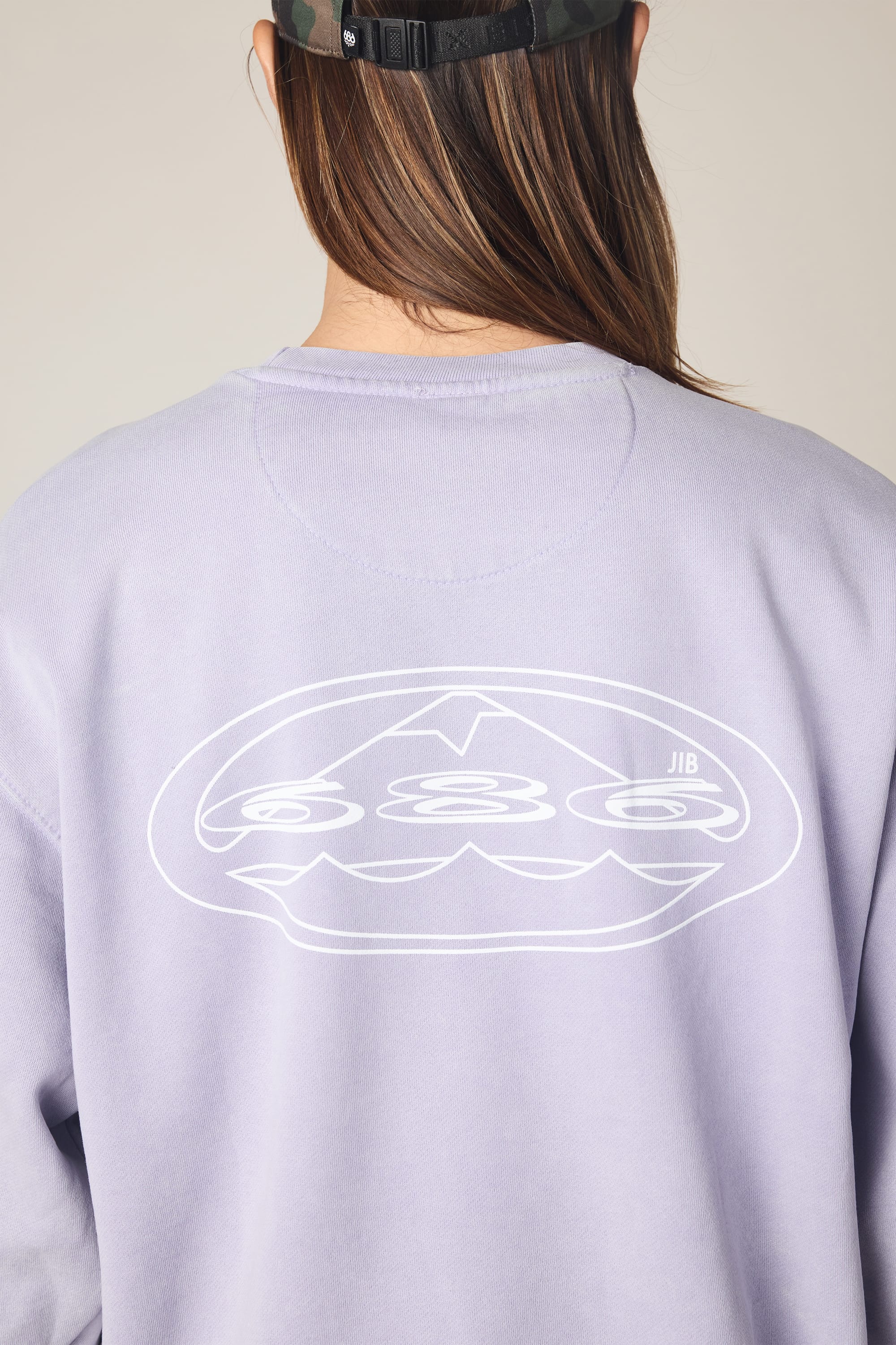 A person with long brown hair wears the 686 Men's Mixed Relaxed LS Crewneck and a black camo-patterned cap turned backwards. The neutral background highlights the 686 brand’s light purple cotton tee with its white abstract back graphic.