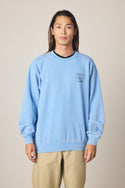 A person with long hair faces forward, wearing the 686 Men's Mixed Relaxed LS Crewneck in light blue with a small left chest graphic, along with beige pants, against a plain, light-colored background.