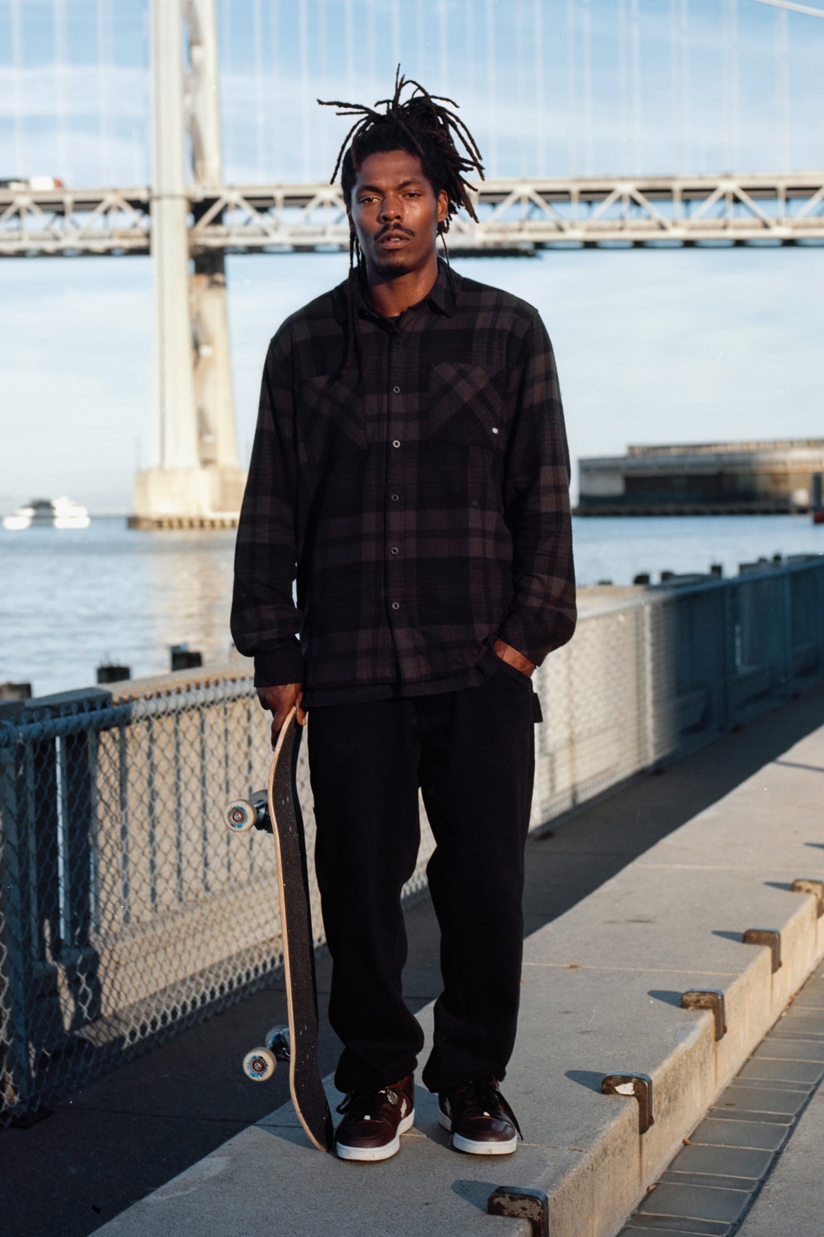 A man standing on a waterfront walkway holding a skateboard, wearing a dark plaid flannel shirt and 686 Men’s Unwork® Hammerhead Denim Pant.